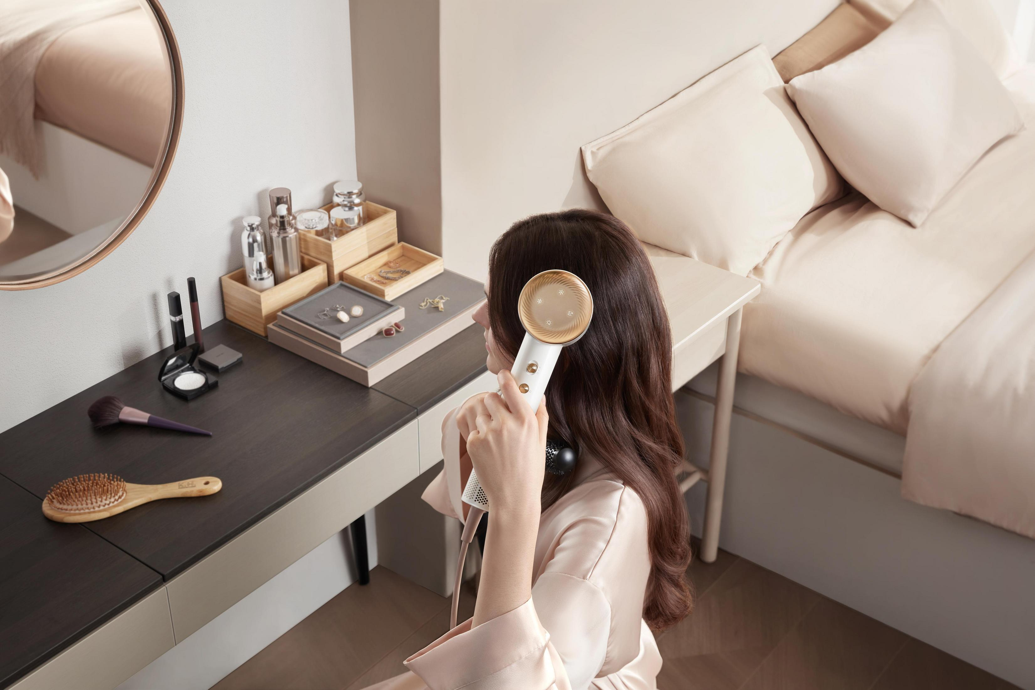 Kobieta używająca narzędzia do stylizacji włosów przy toaletce. Pokój jest beżowy z łóżkiem.