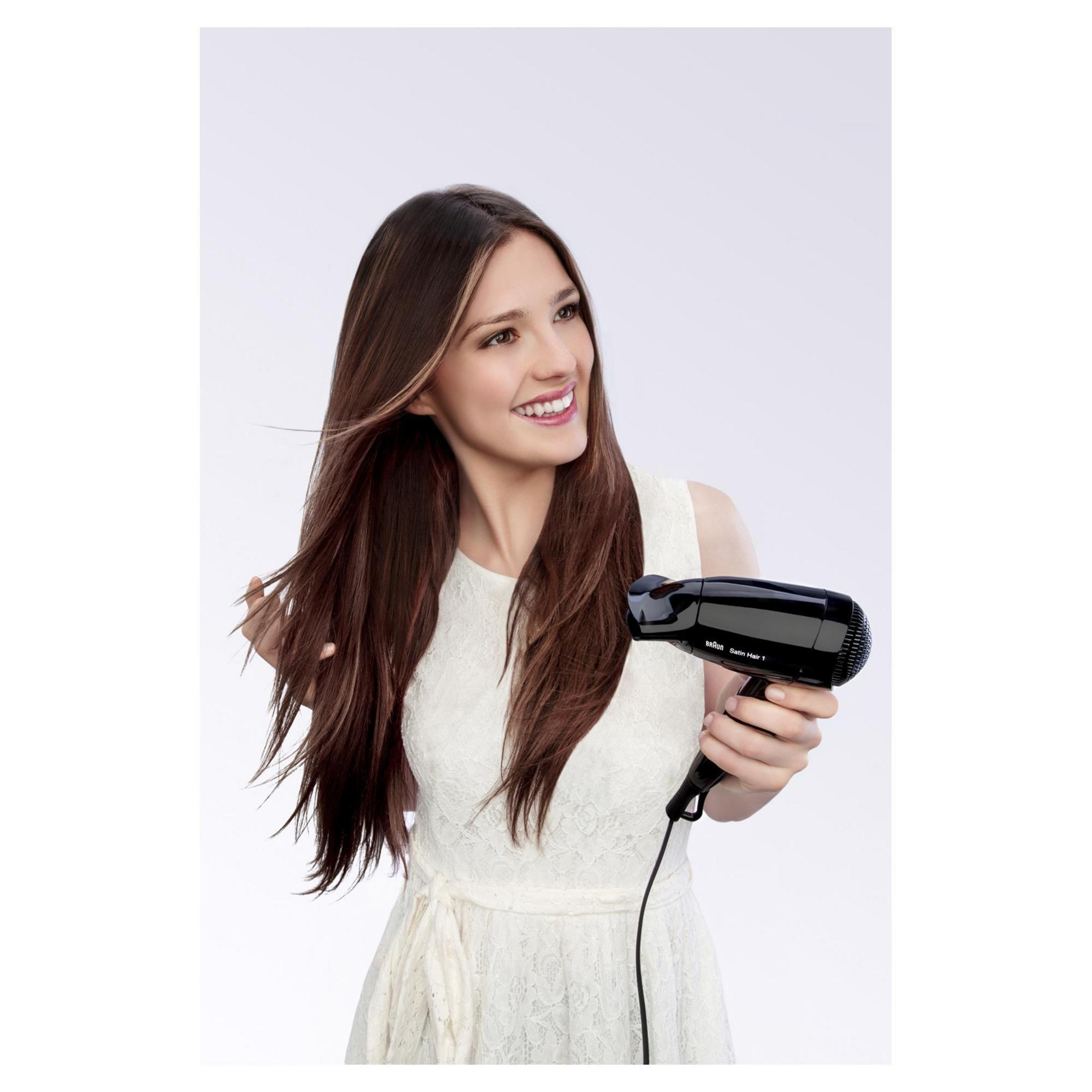 Een vrouw met lang bruin haar lacht terwijl ze een zwarte Braun haardroger vasthoudt, lichte achtergrond.