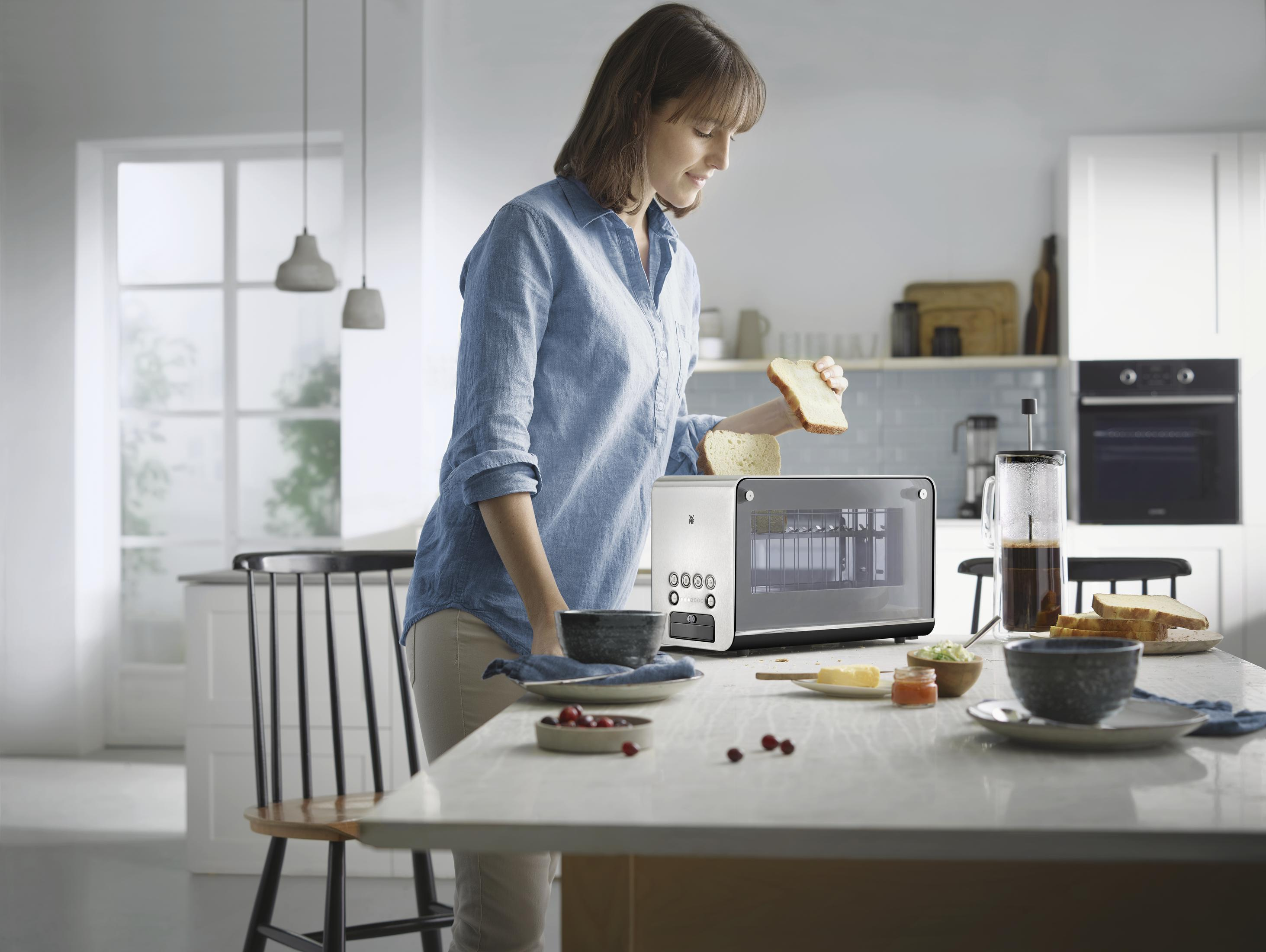 Kobieta w niebieskiej koszuli stoi przy stole i wkłada chleb do tostera.