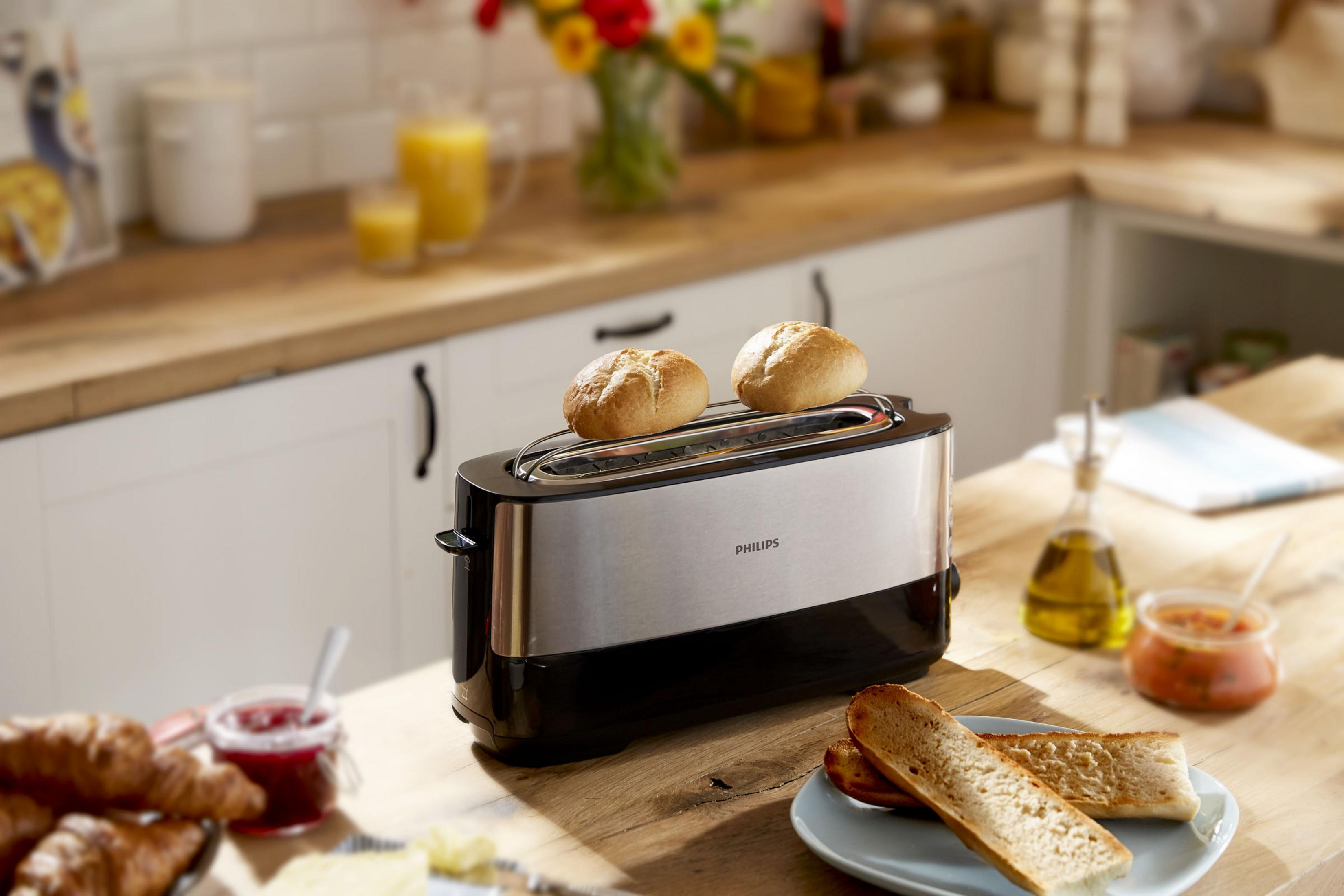 Ein Toaster mit Brot steht auf einem Holztisch. Weitere Lebensmittel sind Toast, Marmelade und Öl. Blumen im Hintergrund.