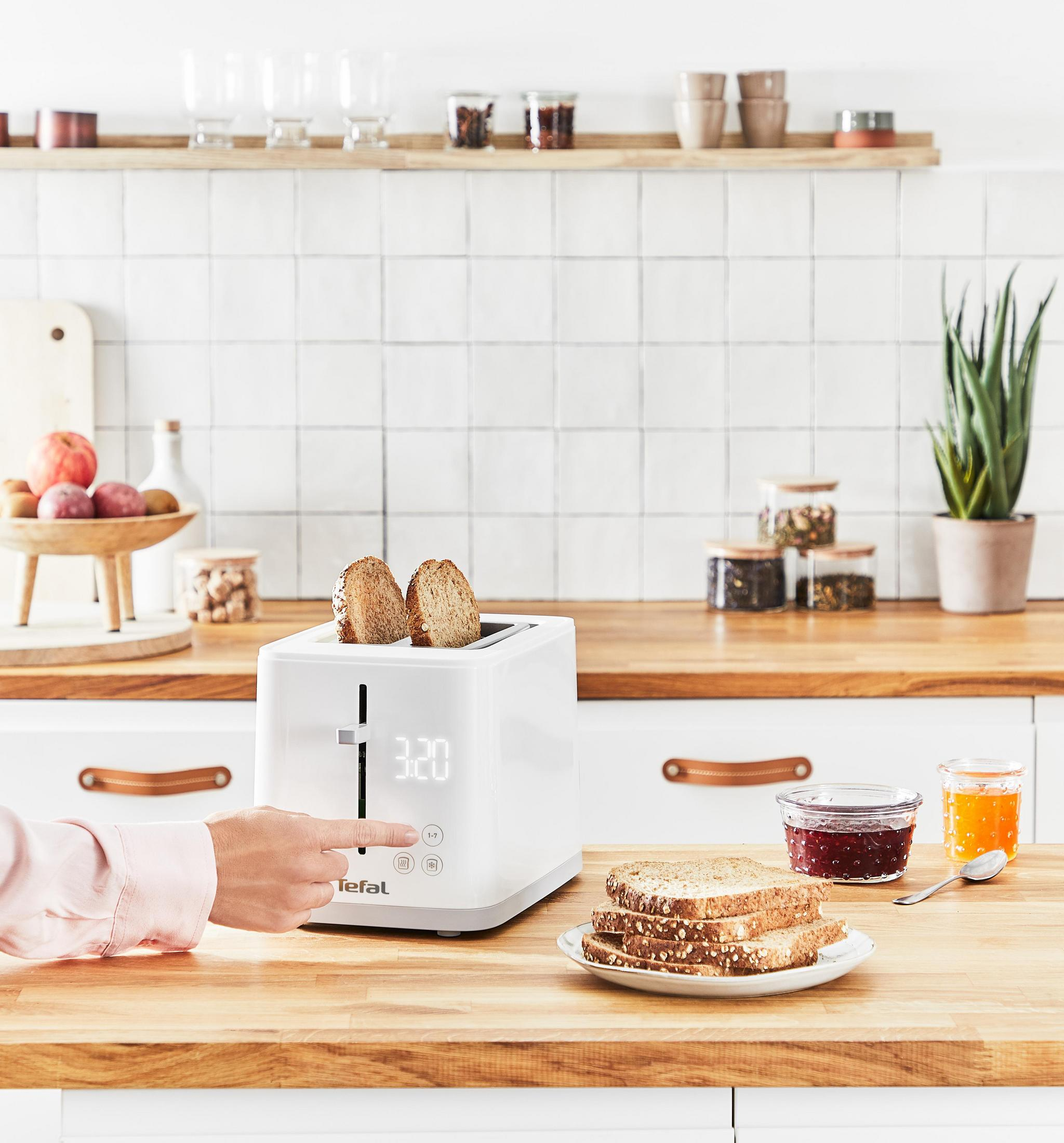 Ein weißer Toaster mit Brotscheiben und Marmeladengläsern auf einer Holz-Arbeitsplatte, mit Hand.