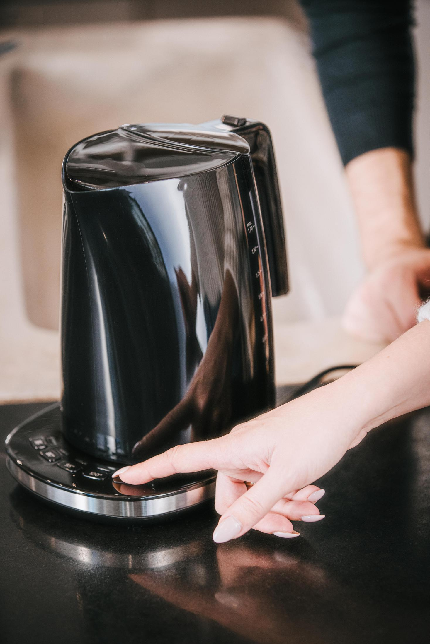 Kobieta naciska przycisk na czarnym czajniku elektrycznym na ciemnej powierzchni.