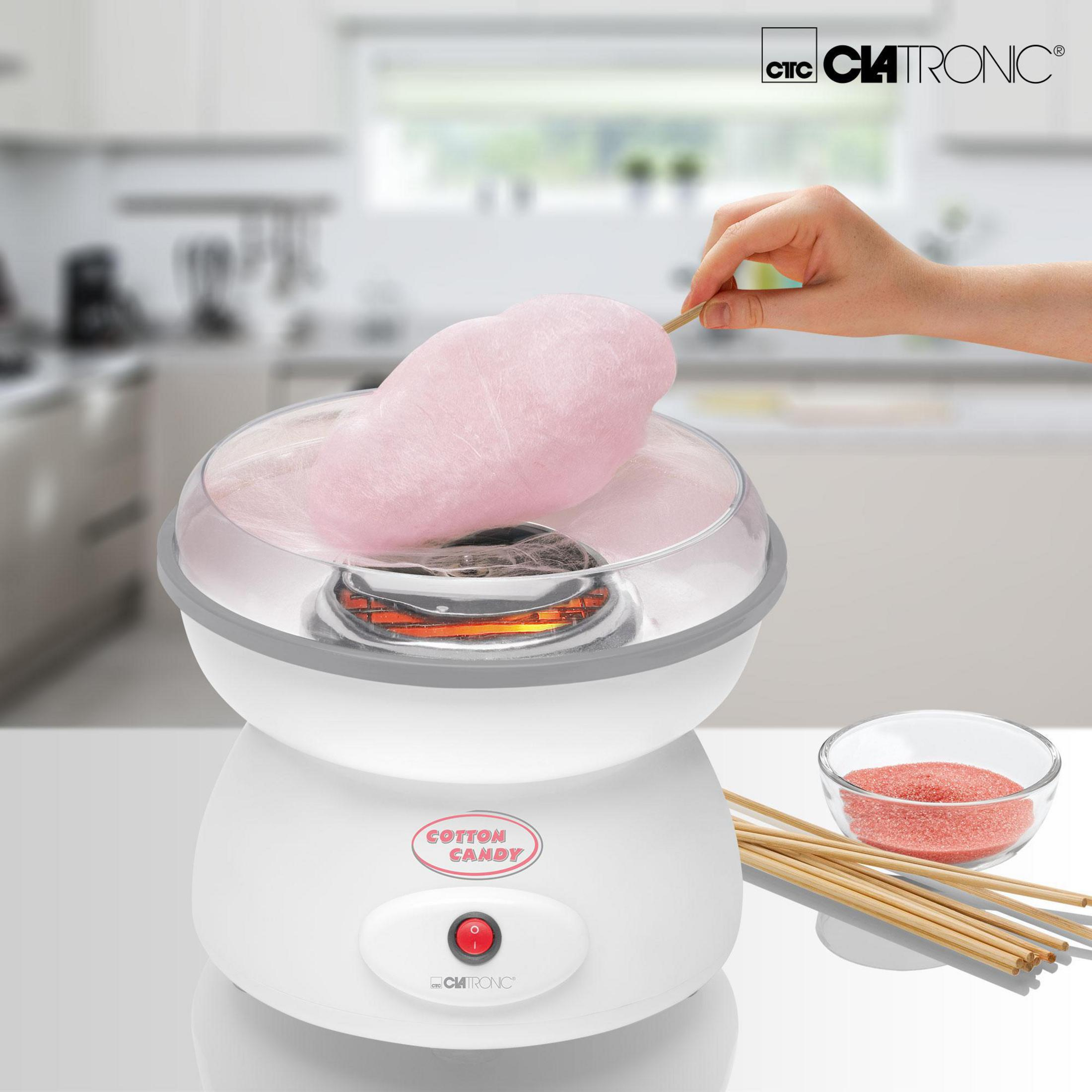 Una persona haciendo algodón de azúcar con una máquina blanca, azúcar rosa y palitos de madera en la mesa.