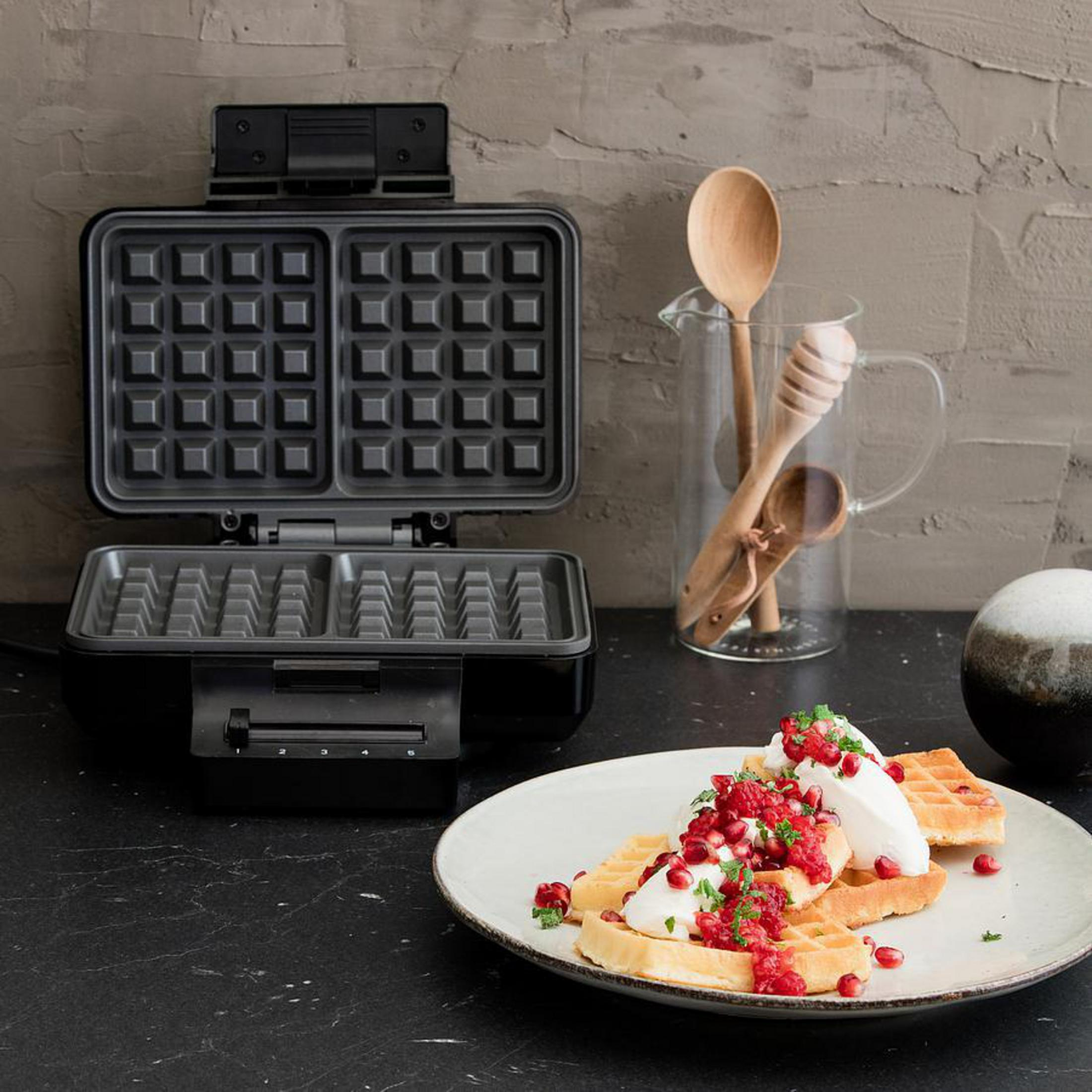 Schwarzer Waffeleisen offen mit Waffeln, Sahne und Beeren auf einem Teller, mit Holzlöffeln.
