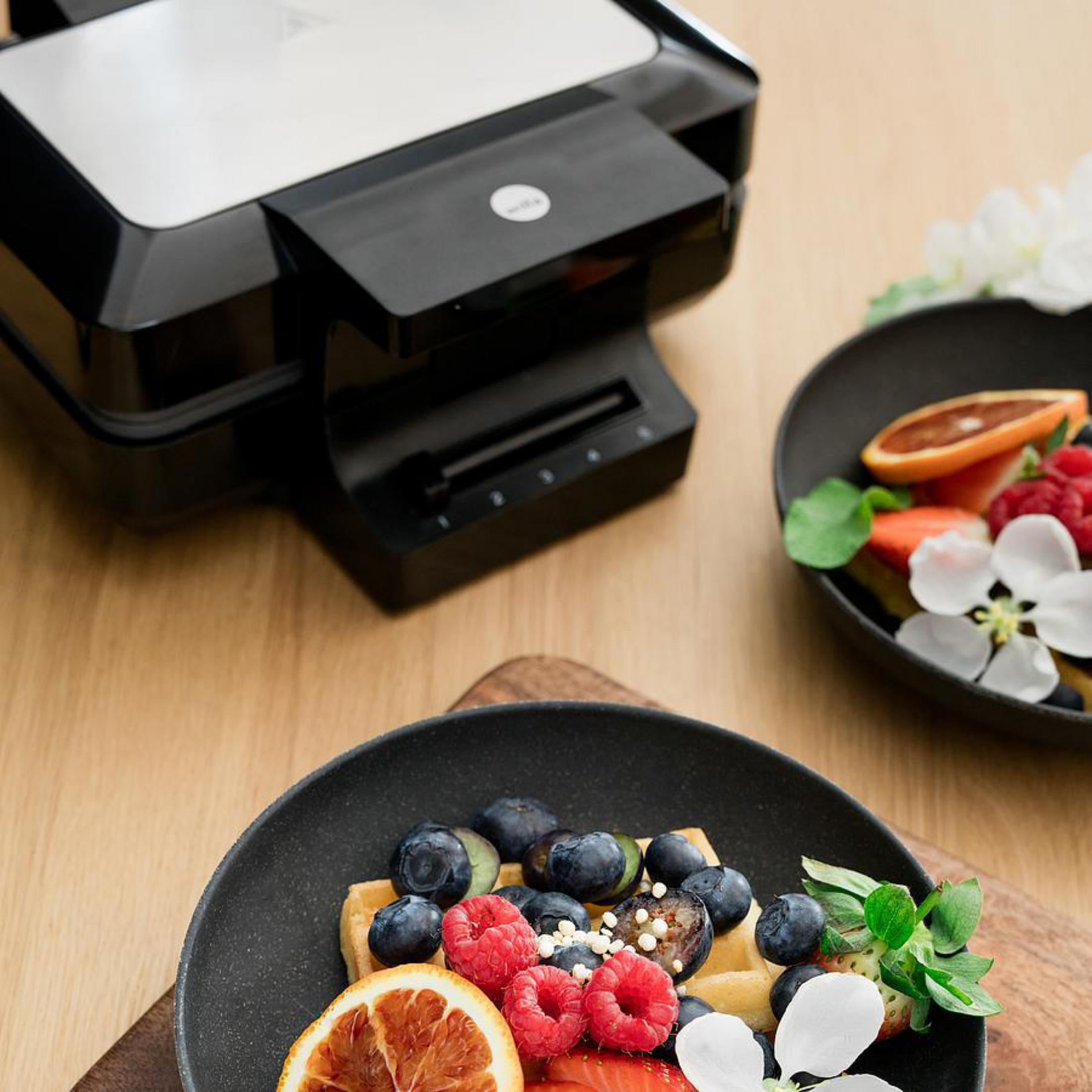 Ein Waffeleisen und ein Teller mit Waffeln, belegt mit Beeren und Fruchtscheiben, auf einem Holztisch.