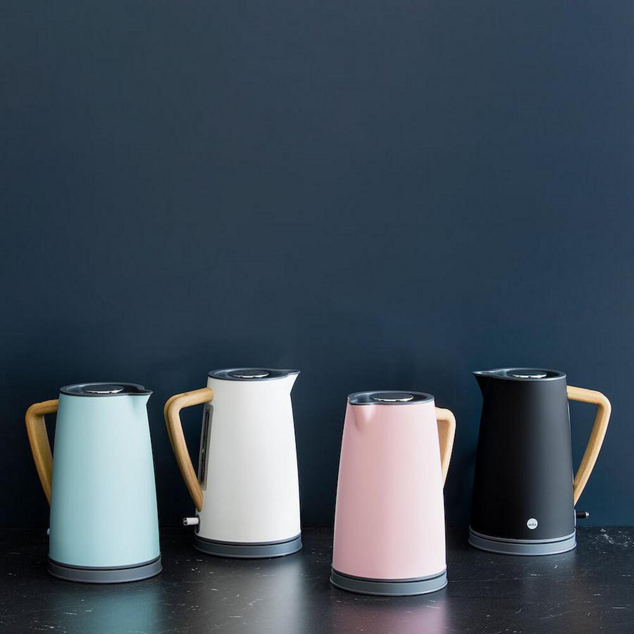 Vier Wasserkocher, hellblau, weiß, rosa und schwarz, mit Holzgriffen, stehen vor einer dunkelblauen Wand.