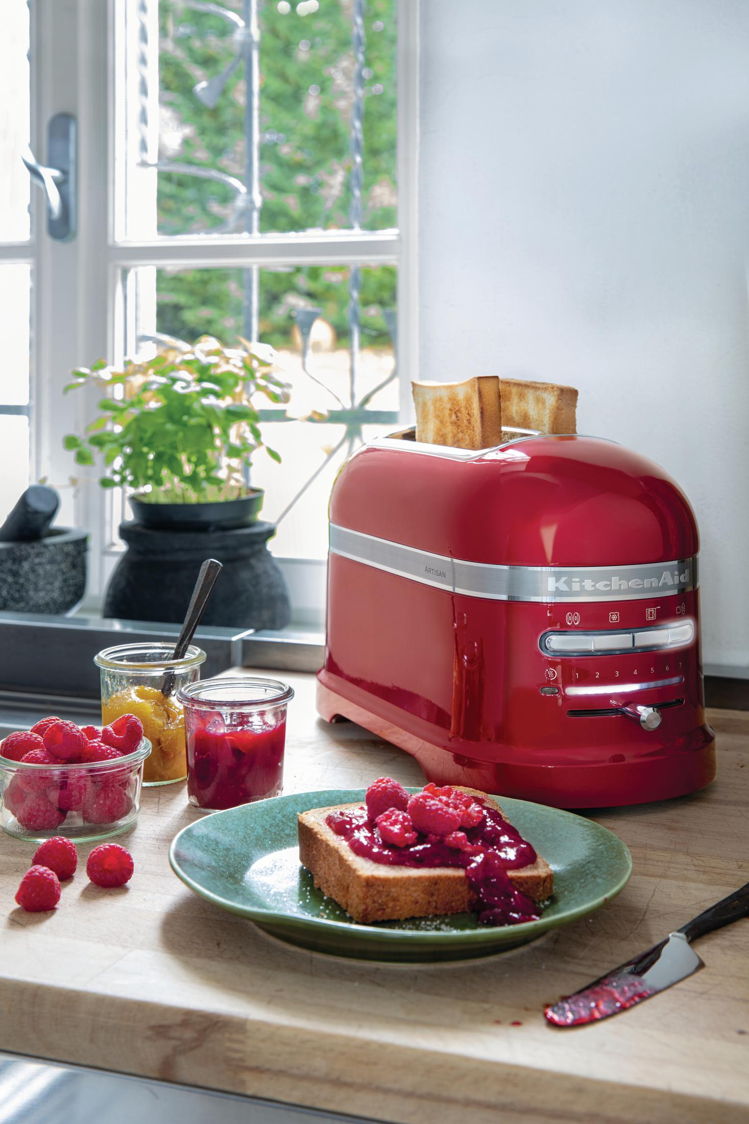 Roter Toaster mit Toast und Marmelade auf einem Teller, Himbeeren und Gläser.