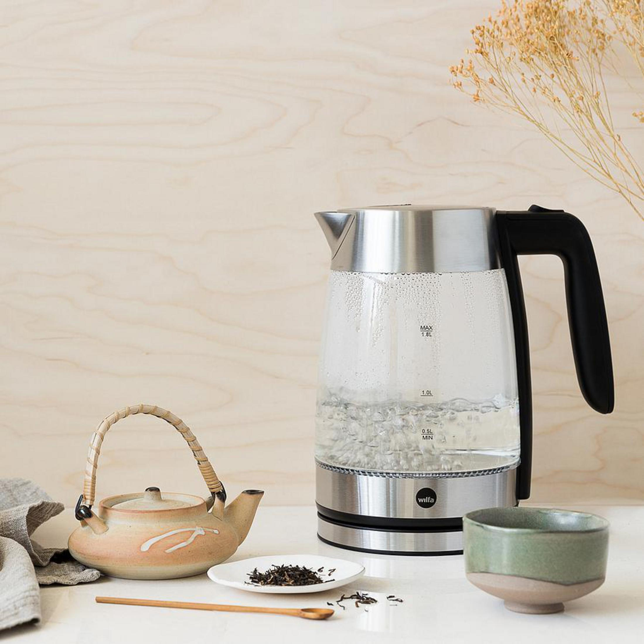 Ein klarer elektrischer Glaskessel mit kochendem Wasser neben einer Teekanne und einer Tasse Tee.