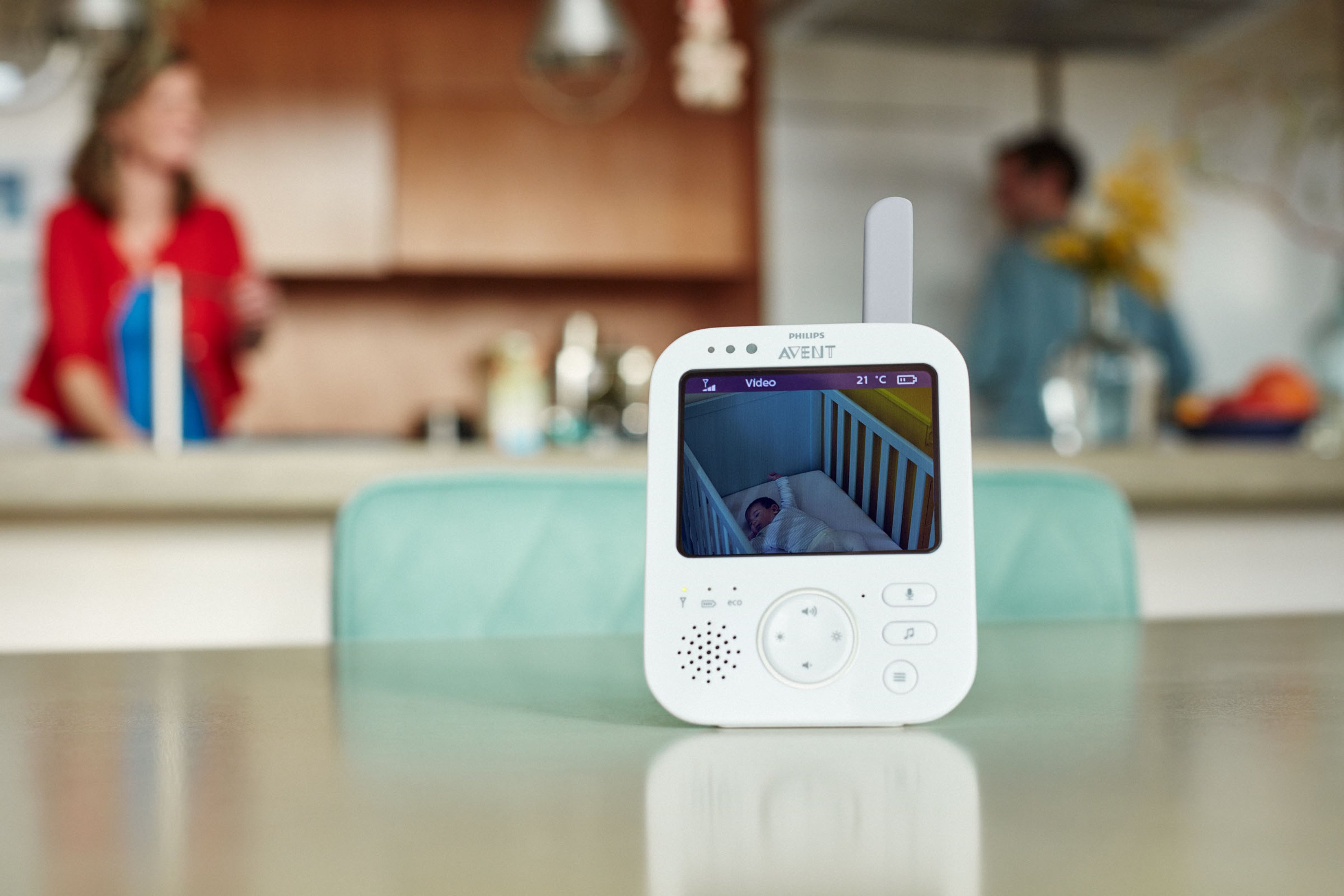 Babyphone, das ein Baby in einer Krippe zeigt, mit Personen im Hintergrund.