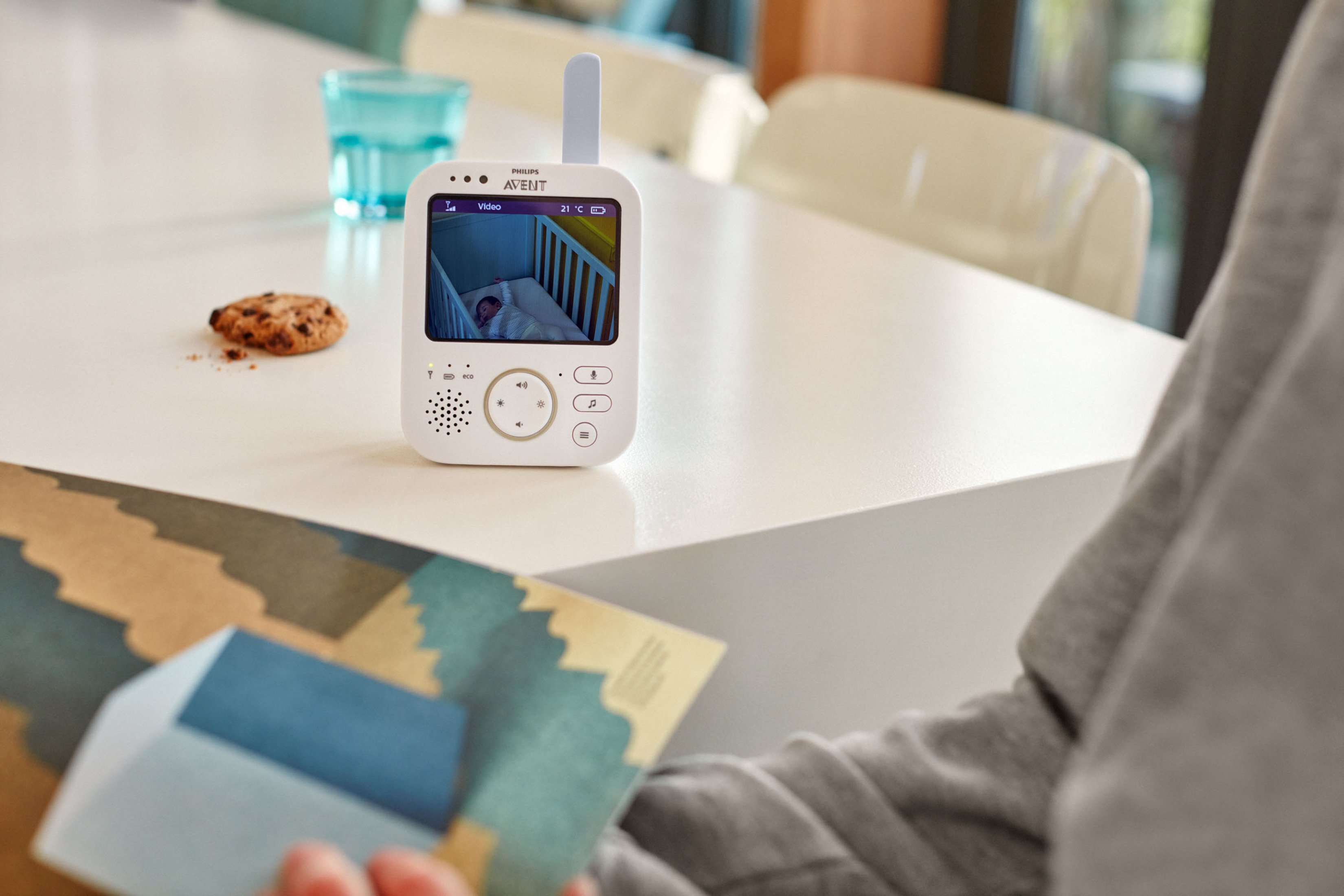 Ein Babyphone zeigt ein Baby im Kinderbett. Eine Person hält eine Karte und ein Keks liegt auf dem Tisch.