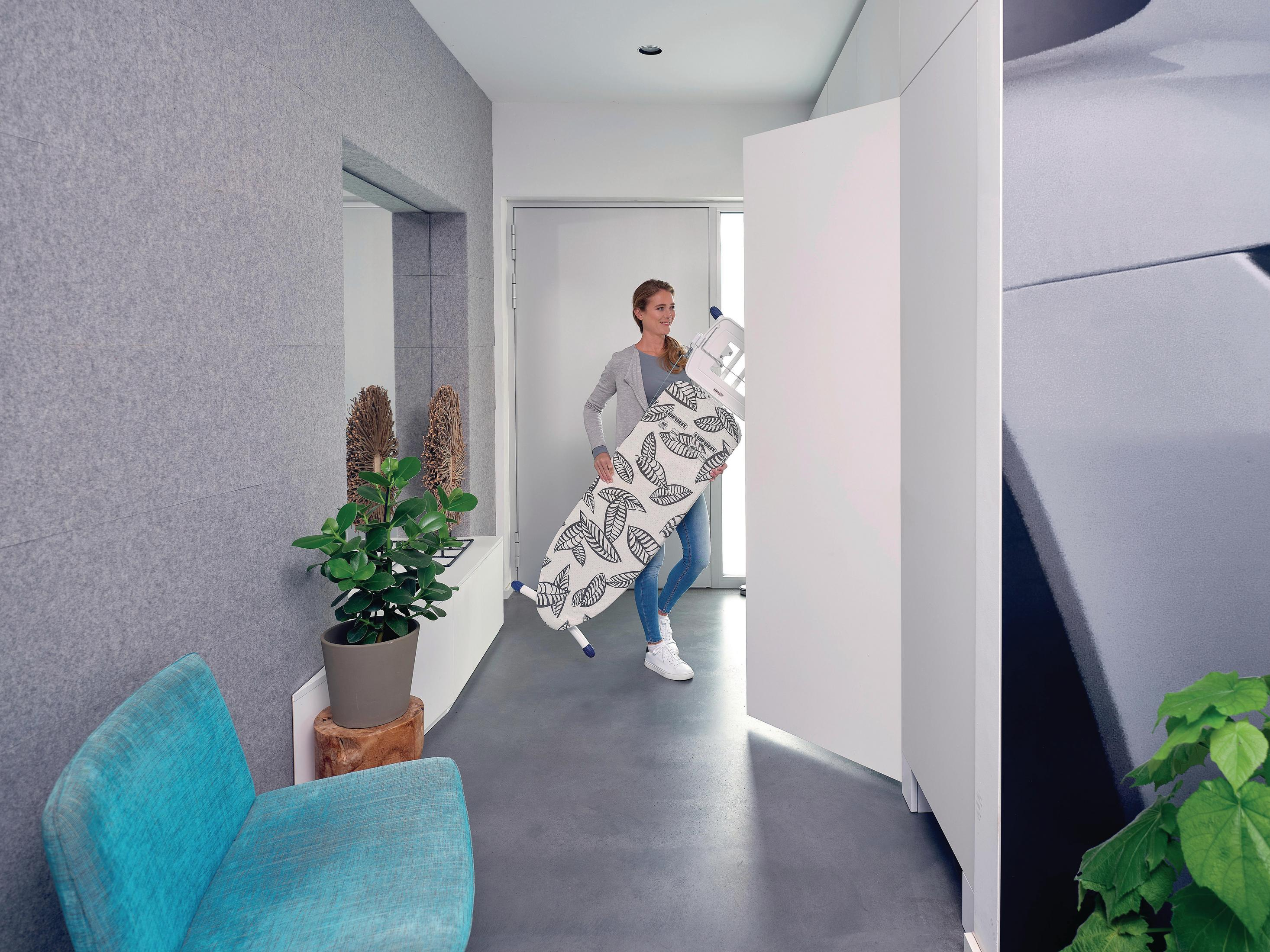 Kobieta niosąca deskę do prasowania we wzór liści przez drzwi, tło korytarza.