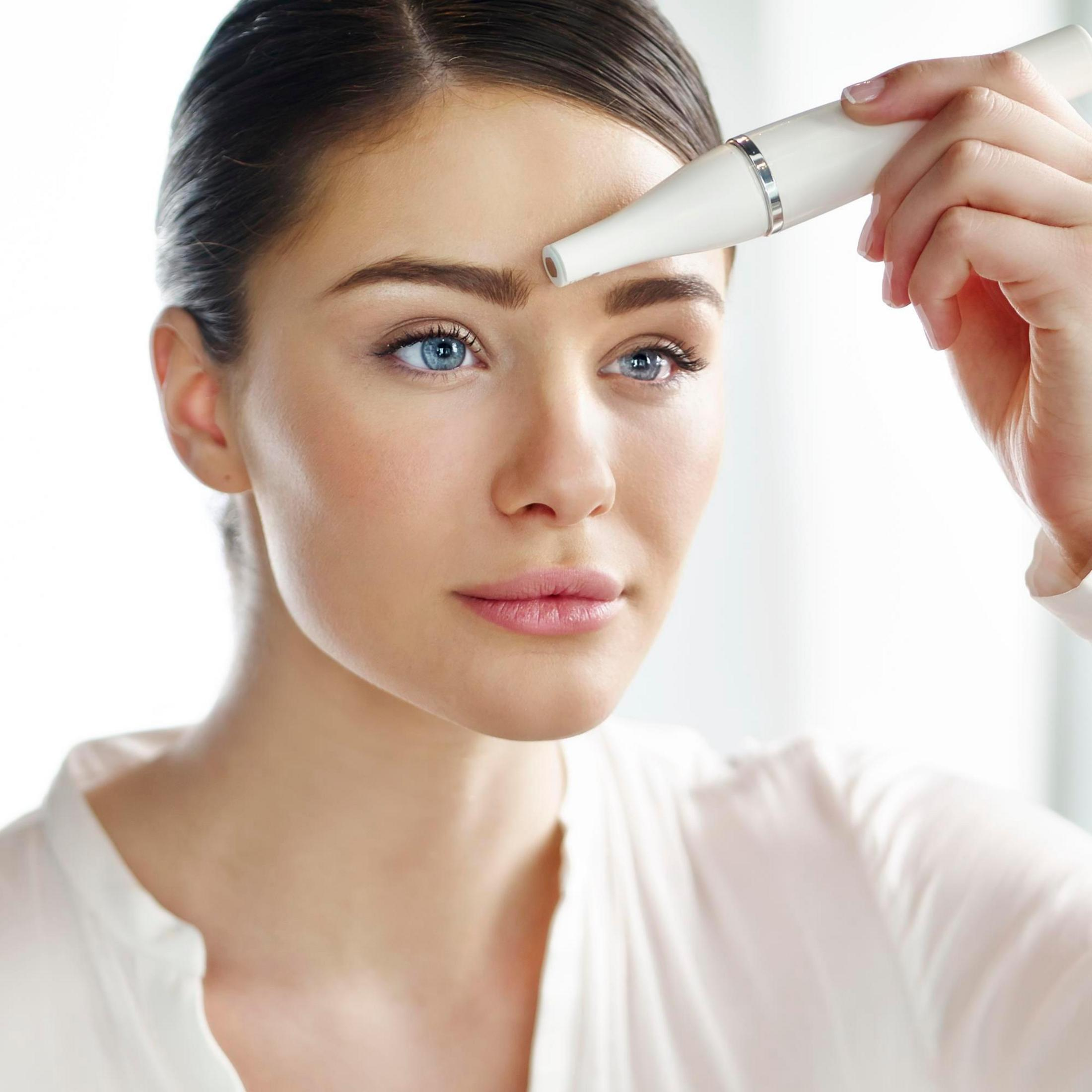 Kobieta używająca trymera do brwi, zbliżenie na jej twarz z niebieskimi oczami i jasną skórą.