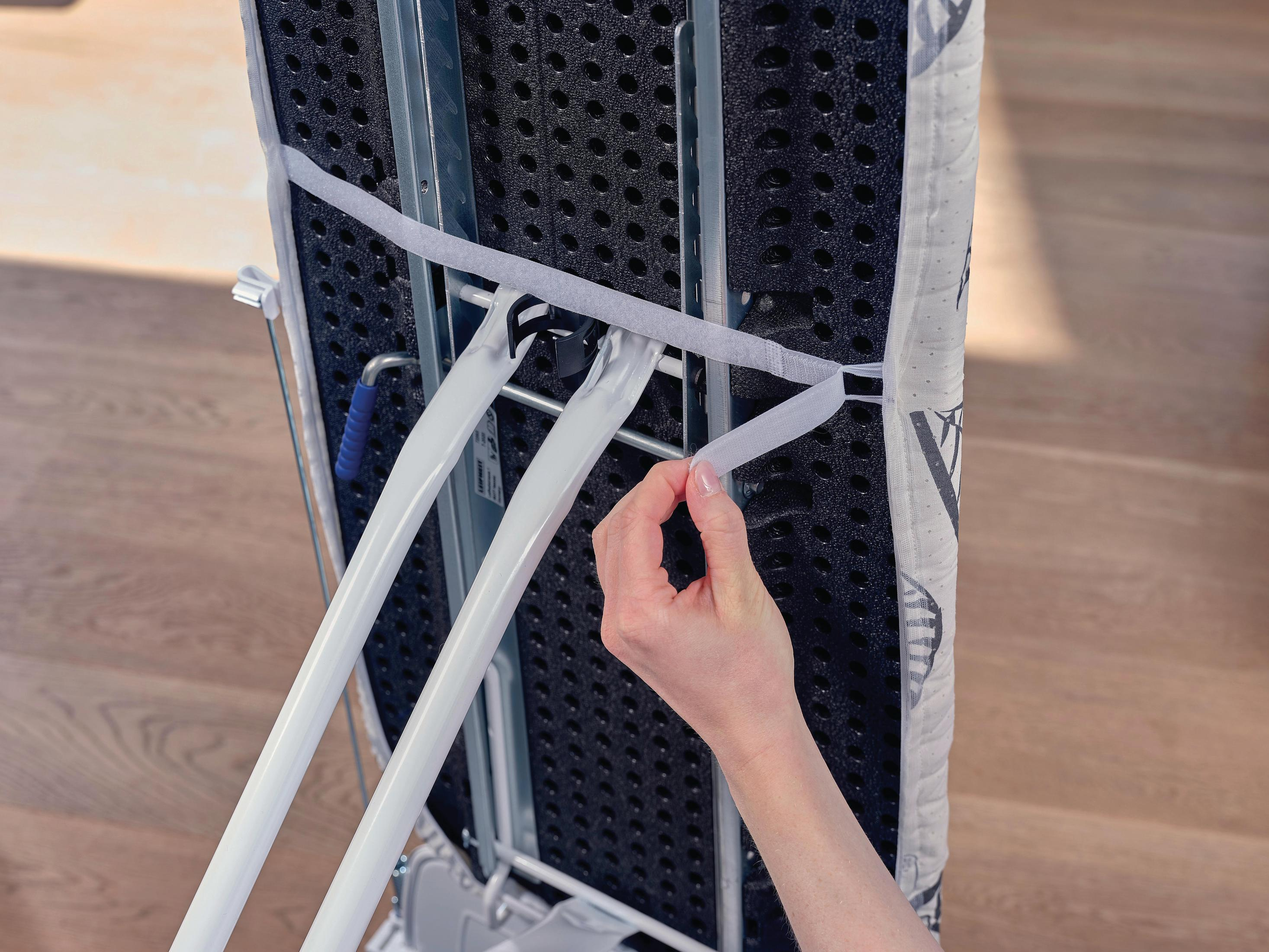 Ręka zapina pasek na rzep na pokrowcu deski do prasowania, z jasną drewnianą podłogą.