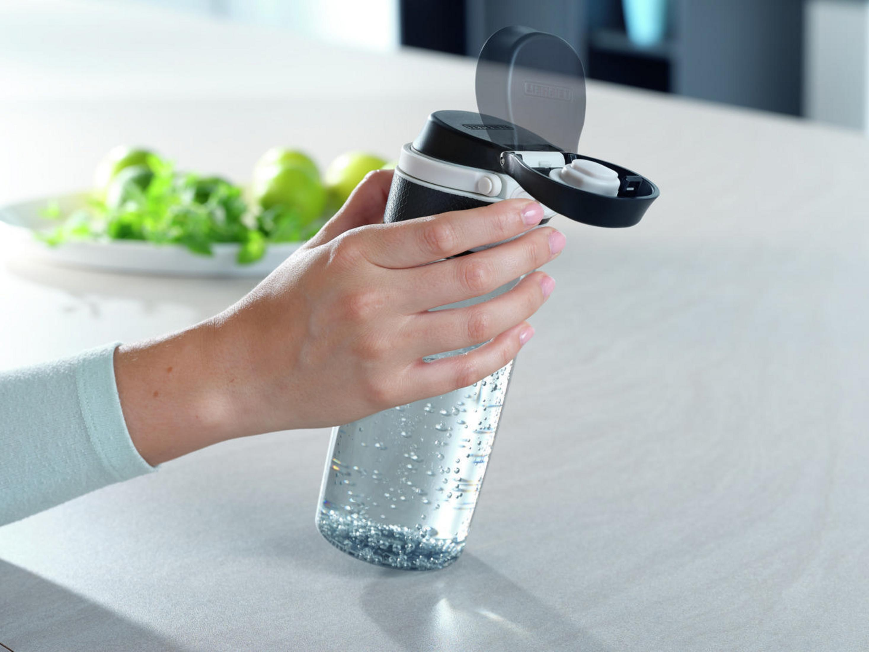 Mujer sosteniendo una botella de agua, tapa abierta, con vegetación en el fondo.