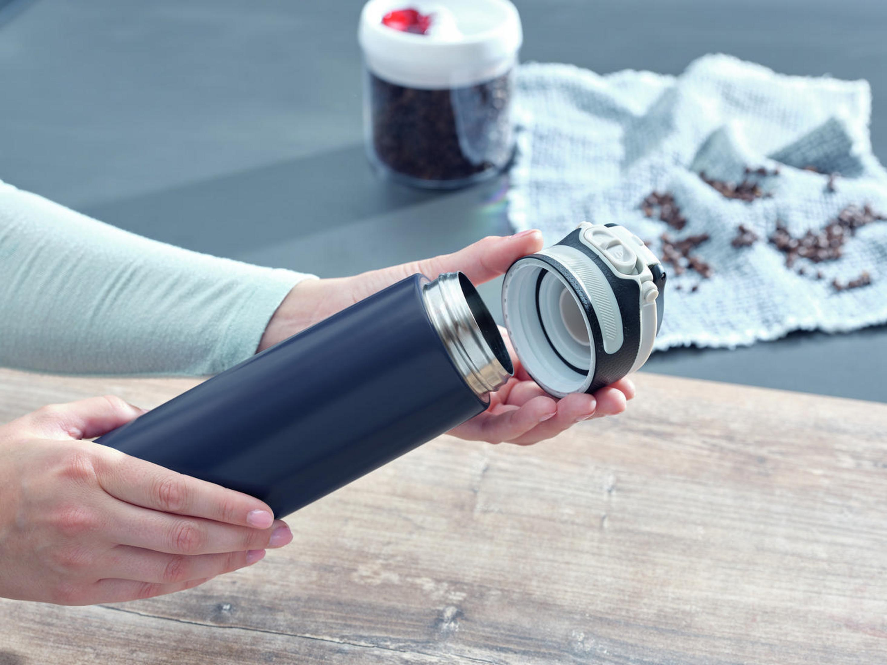 Person hält eine dunkelblaue Thermoskanne und schraubt den Deckel ab. Kaffeebohnen sind sichtbar.