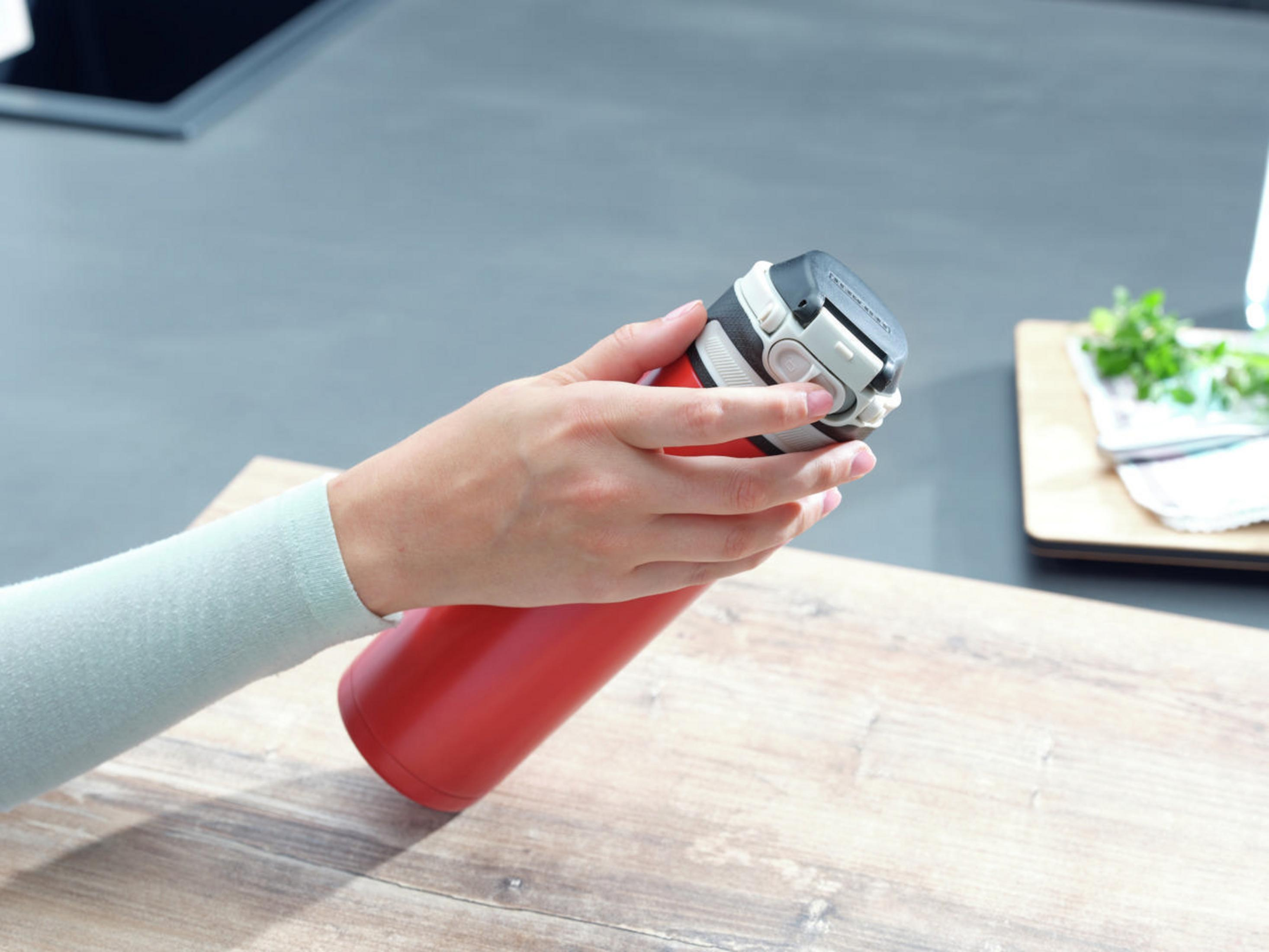 Eine Hand hält eine rote Thermosflasche, der Deckel ist geöffnet. Auf einer Holzoberfläche.