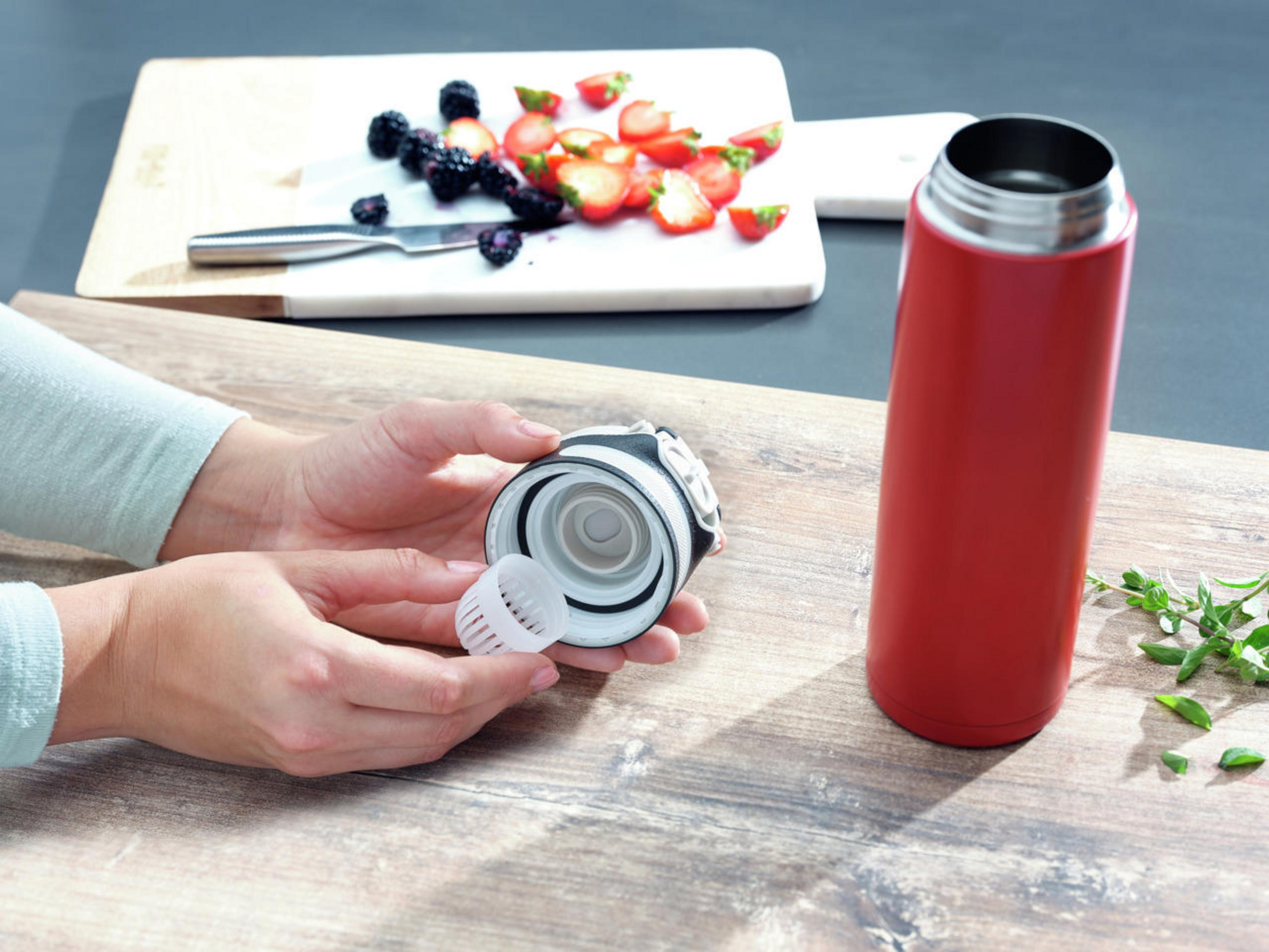 Person hält eine rote Thermosflasche, Deckel offen, Filter sichtbar. Beeren und Erdbeeren daneben.