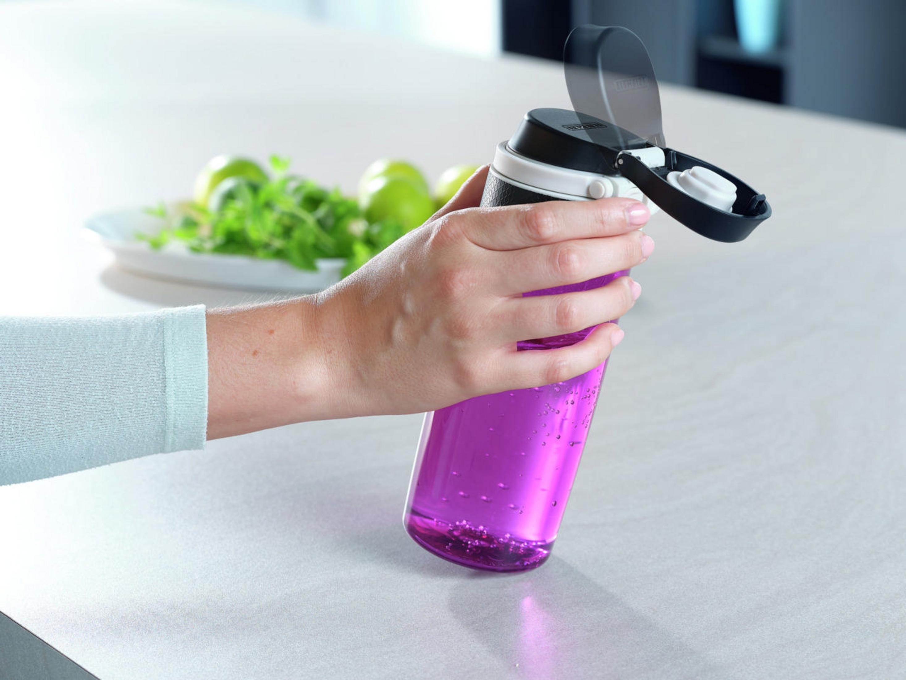 Una mano sostiene una botella de agua morada con la tapa abierta. Limas verdes están en el fondo.