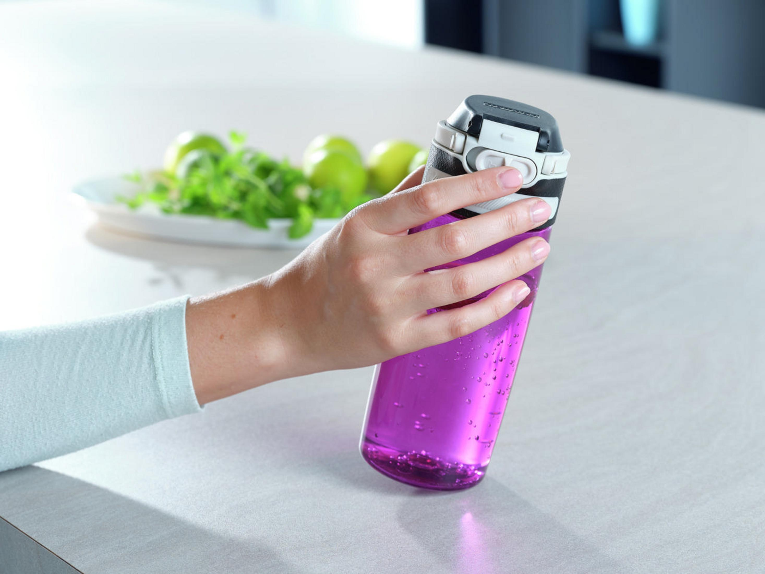 Una persona sostiene una botella de agua morada. Manzanas verdes y hierbas en un plato blanco al fondo.