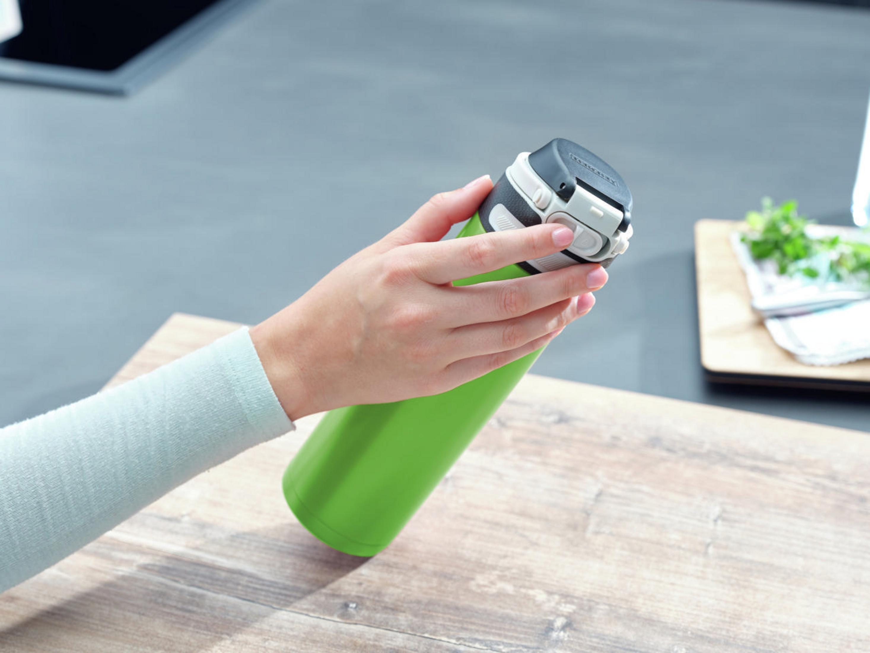 Hand hält grüne Flasche, im Hintergrund grauer Tisch.