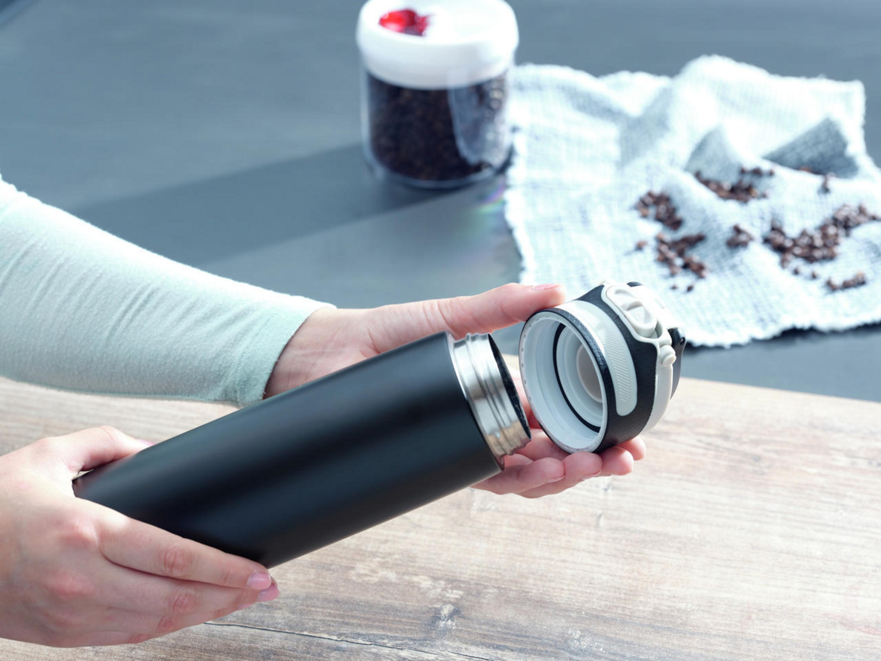 Hände öffnen eine schwarze, isolierte Wasserflasche. Ein Behälter mit Kaffeebohnen und ein Tuch sind ebenfalls sichtbar.