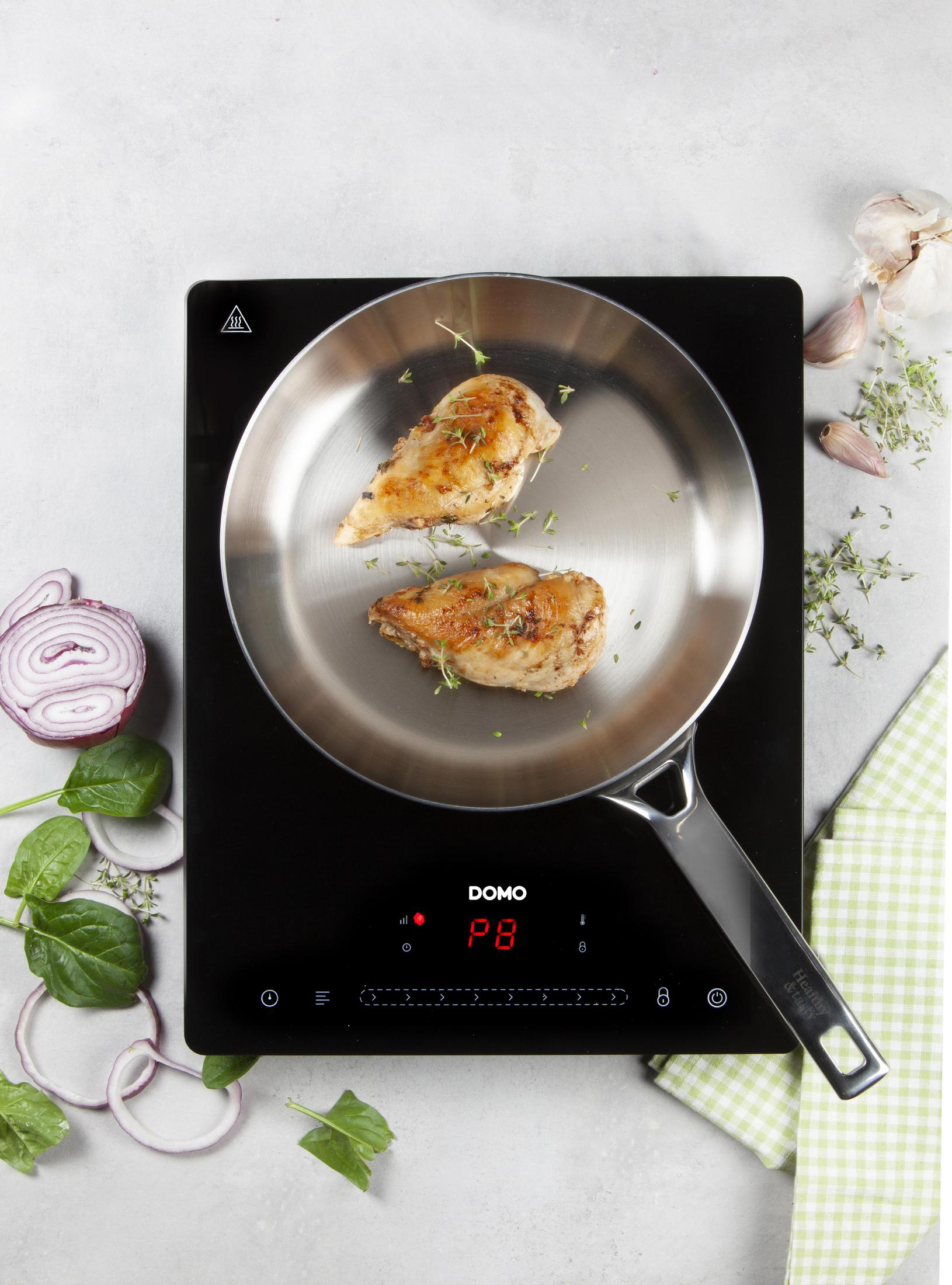 Dwie piersi z kurczaka w patelni na czarnej płycie indukcyjnej, z ziołami i warzywami w pobliżu.