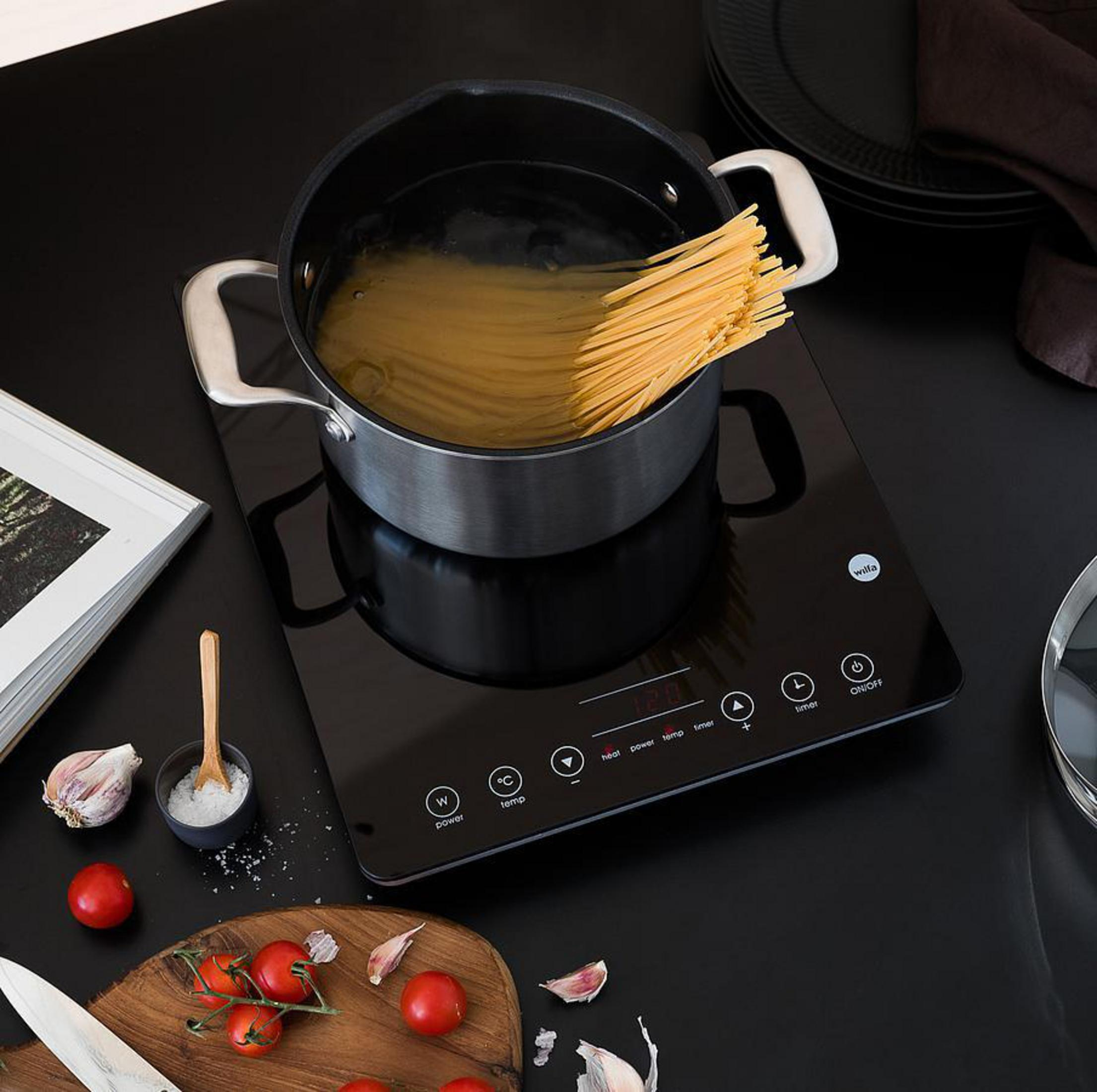 Placa de inducción negra con espaguetis en una olla. Ajo, tomates y sal.