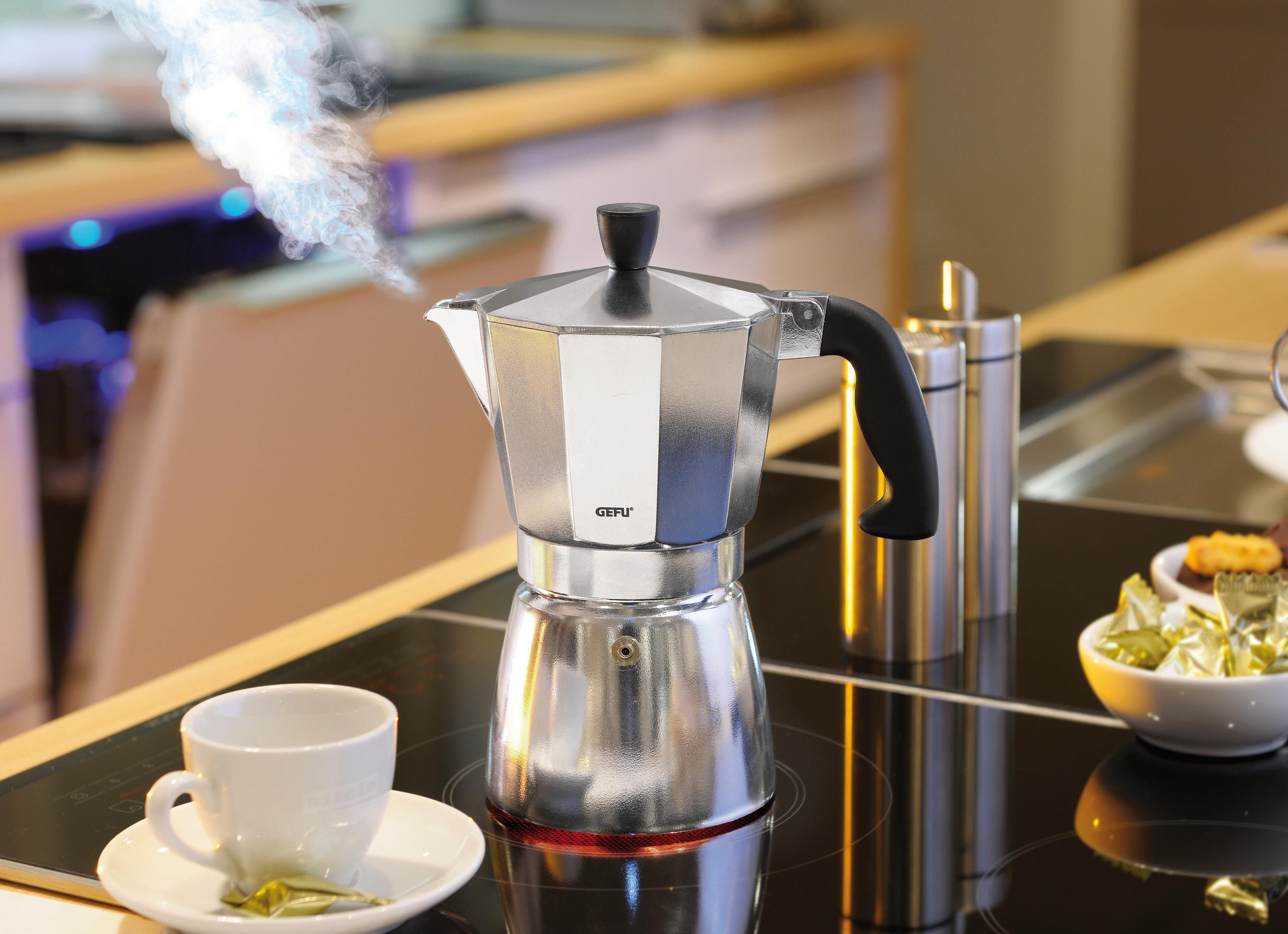 Kaffeemaschine mit Dampf, Tasse und Untertasse auf schwarzem Herd, unscharfer Hintergrund.