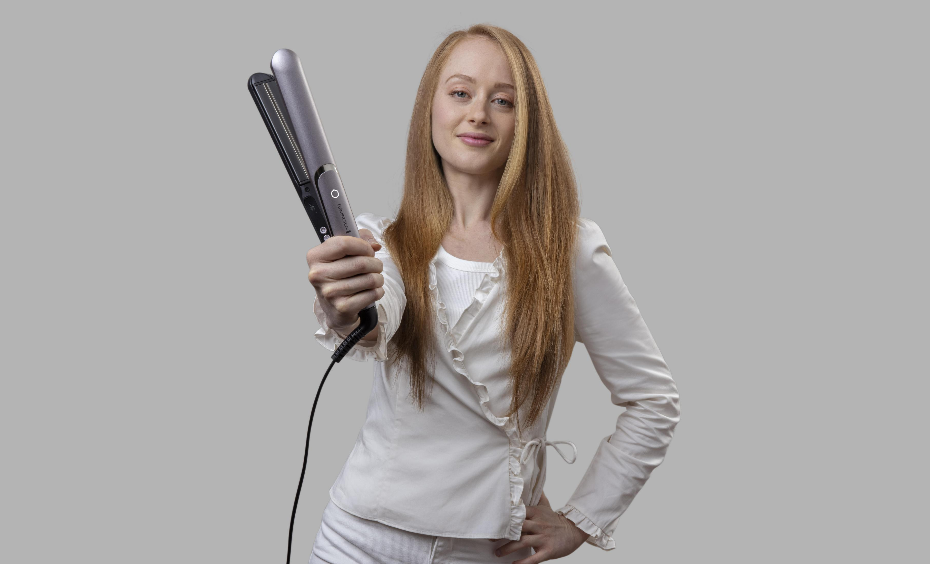 Una donna con i capelli rossi mostra una piastra per capelli, sorridendo. Indossa abiti bianchi su sfondo grigio.