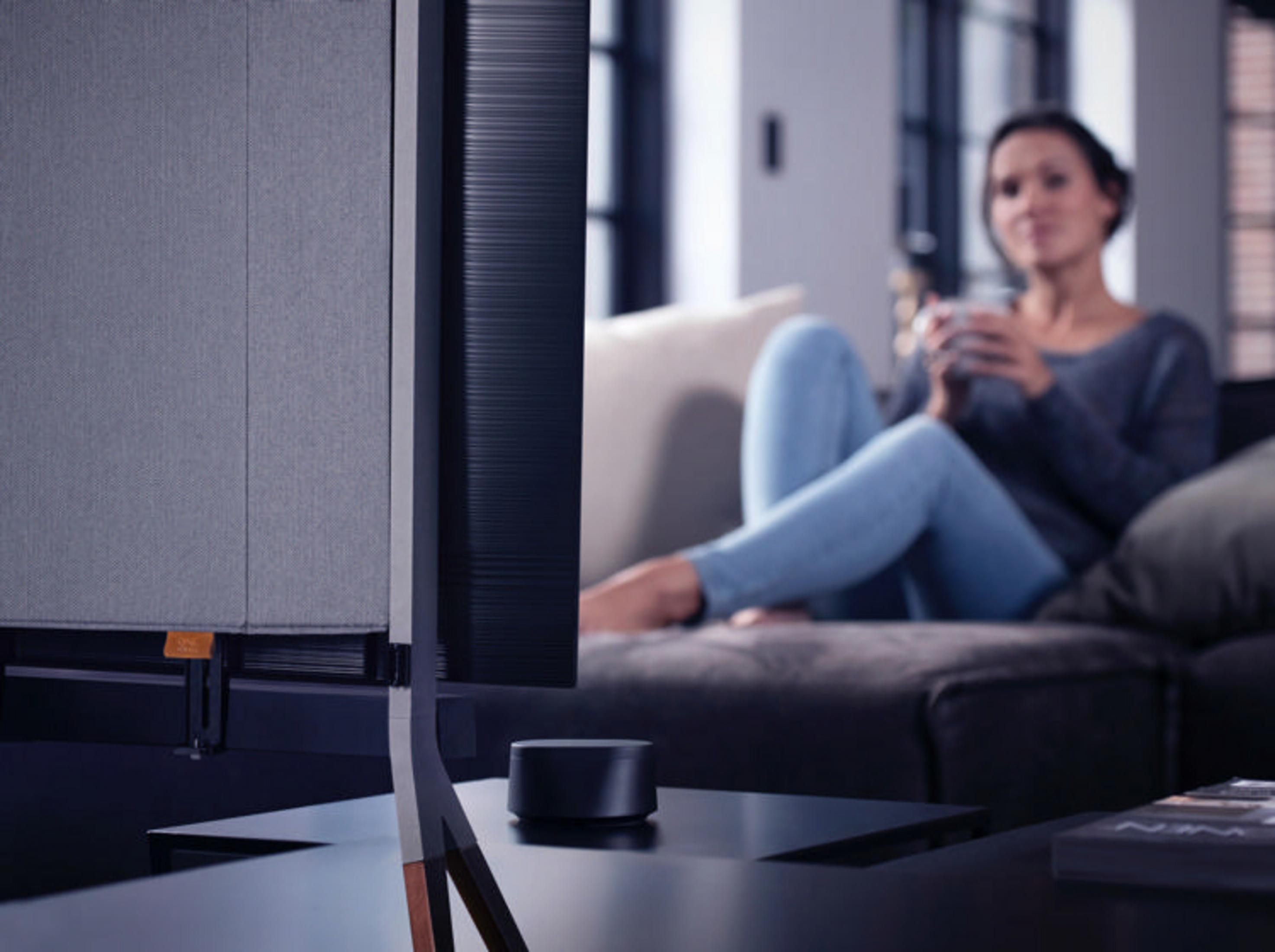 Televisore moderno con pannello in tessuto. Una donna è su un divano sullo sfondo. Una soundbar nera è su un tavolo.