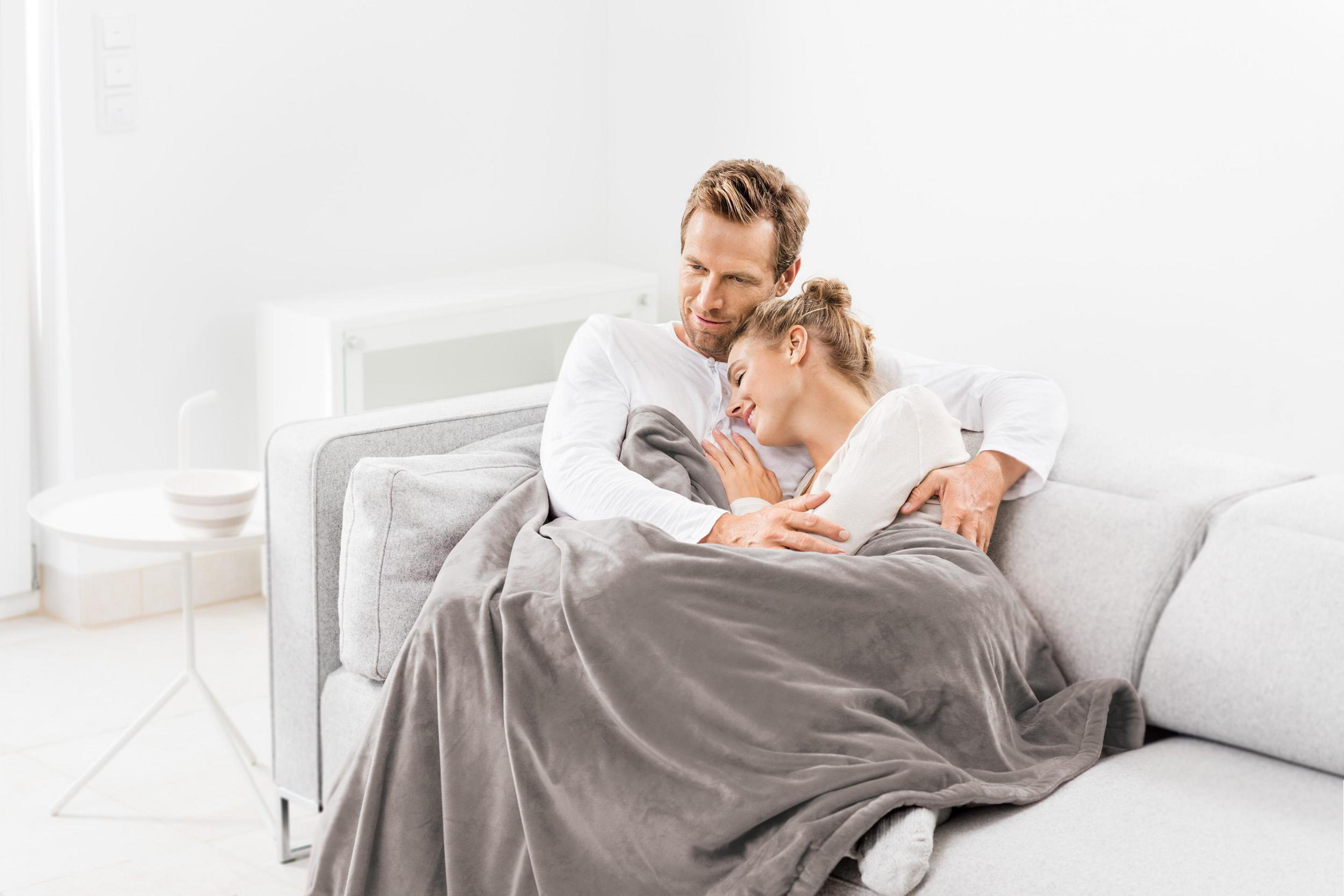 A man and a woman wrapped in a blanket, sitting together on a light gray sofa.