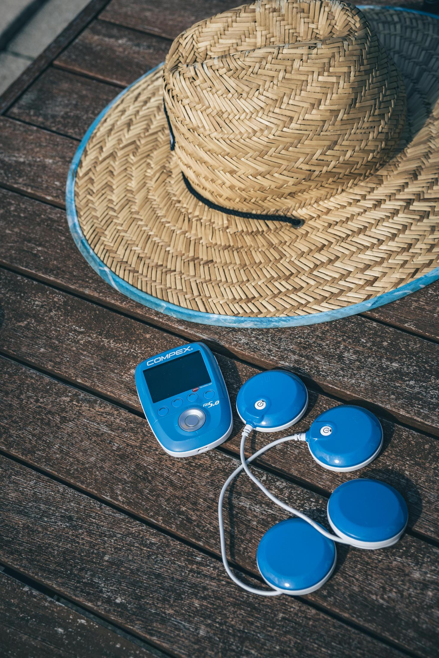 Strohhut und blaues Compex-Gerät mit blauen Pads auf Holz, sonniger Tag.