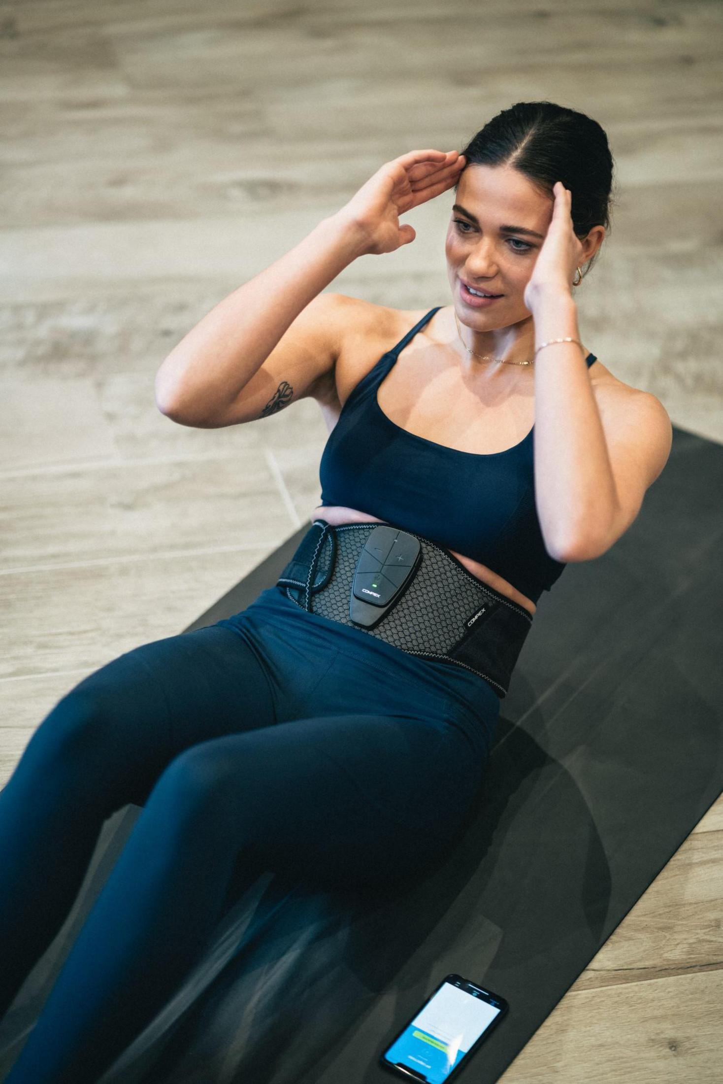 Frau mit Fitnessgürtel beim Sit-ups auf einer schwarzen Matte, Telefon zur Seite.