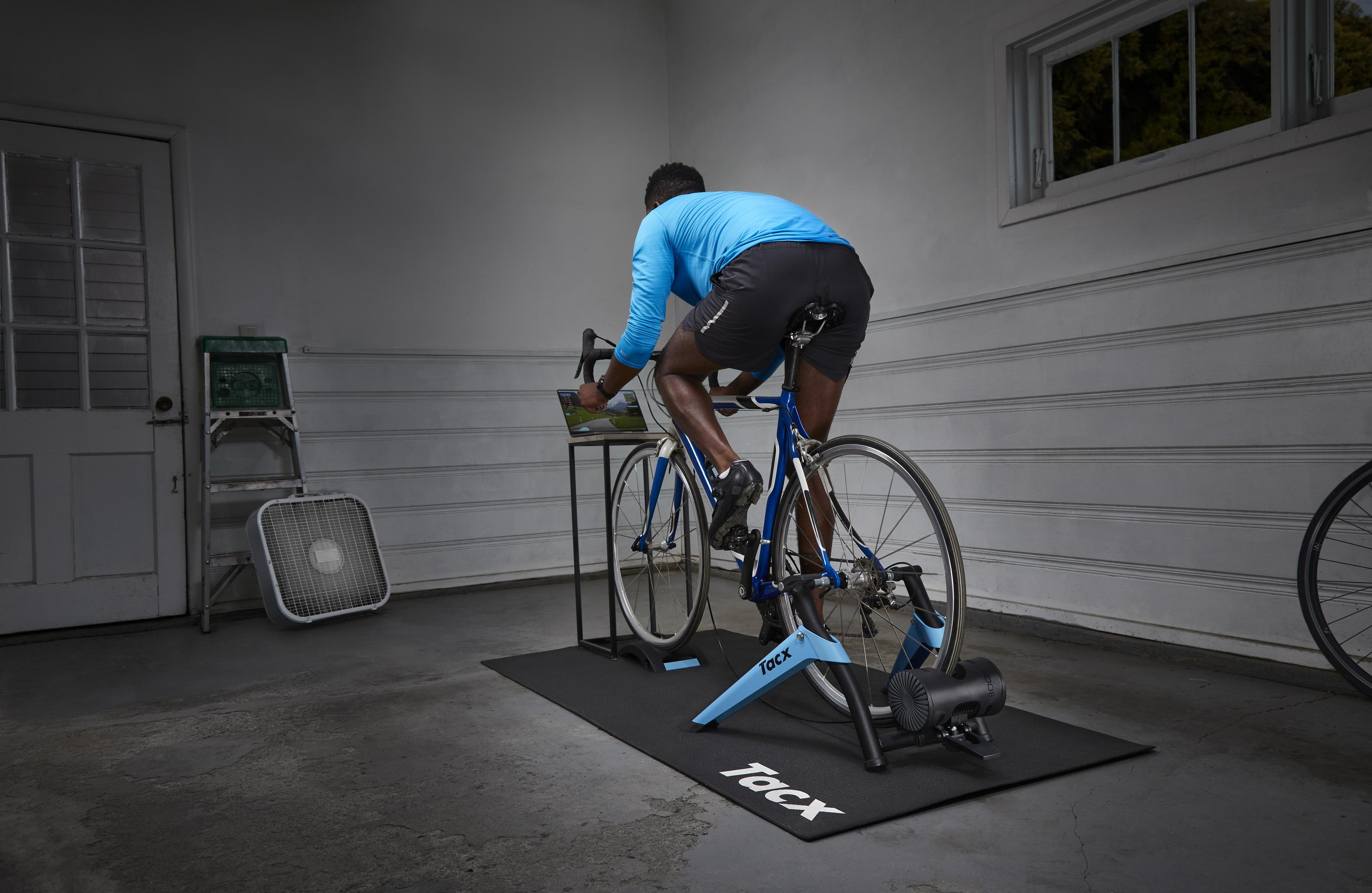 Ein Mann fährt auf einem blauen Fahrrad auf einem Trainer in einer Garage. Ein Ventilator und eine Kiste sind links.