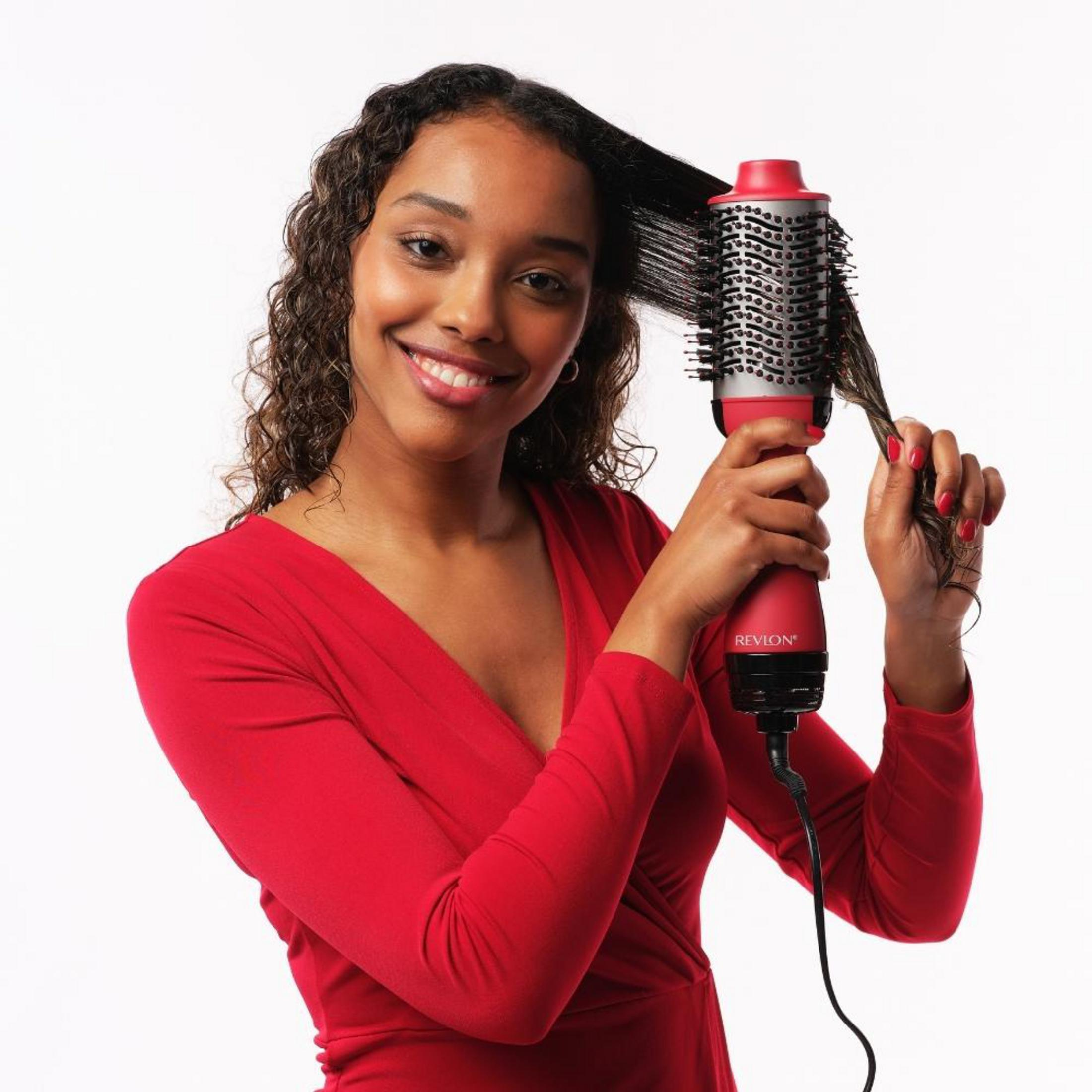 Una donna che si acconcia i capelli con una spazzola ad aria calda rossa, sorridendo alla fotocamera.