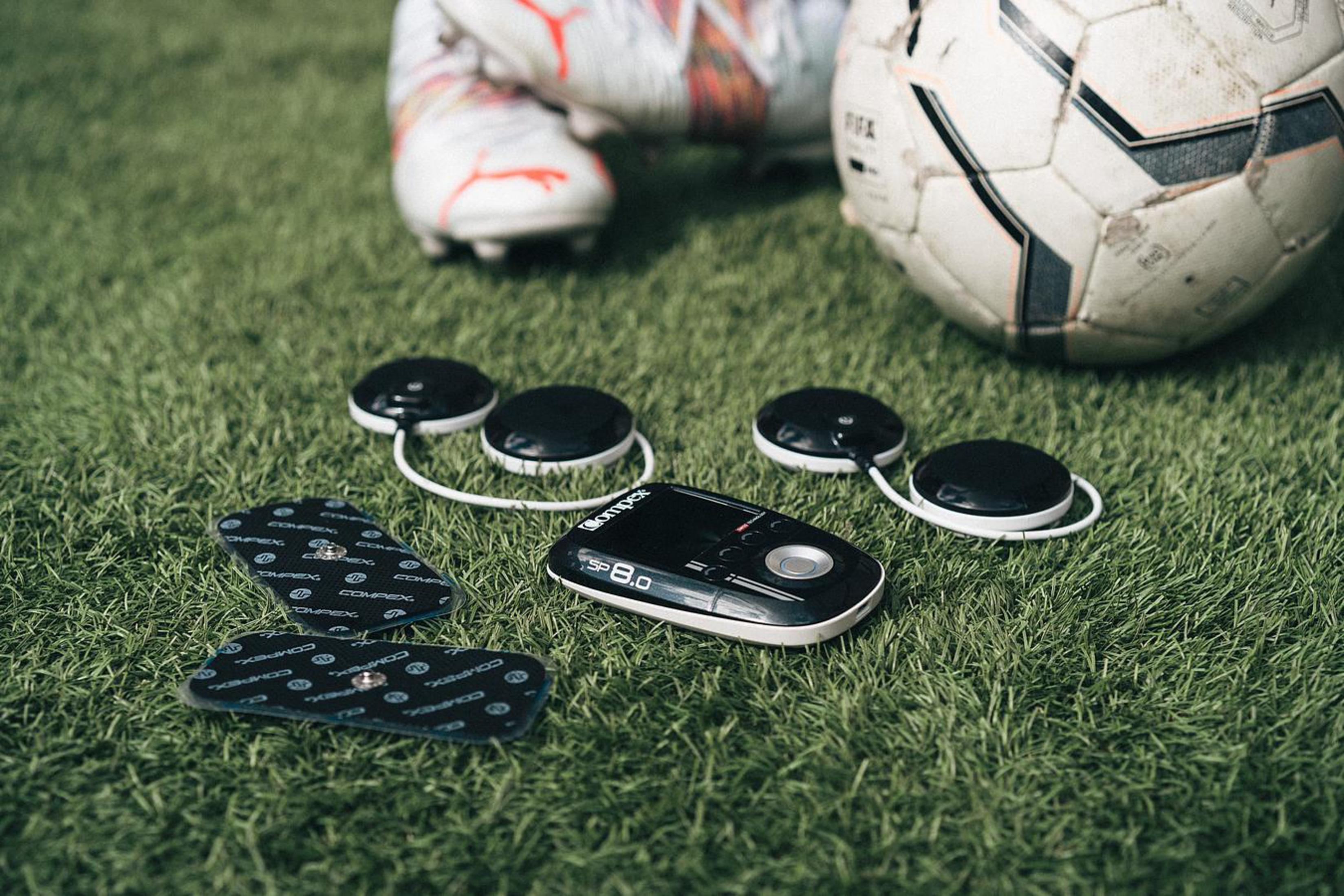 Fußball, Schuhe und ein Compex-Gerät auf grünem Rasen.