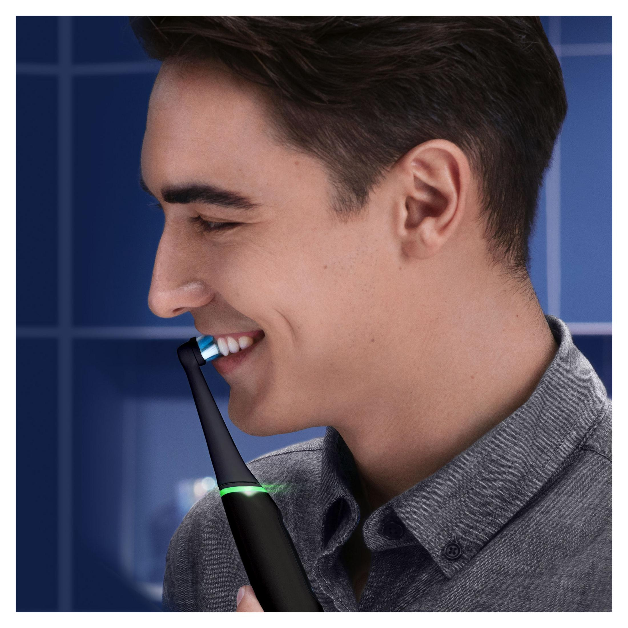 Mężczyzna używający czarnej elektrycznej szczoteczki do zębów Oral-B, uśmiechnięty. Niebieskie tło.