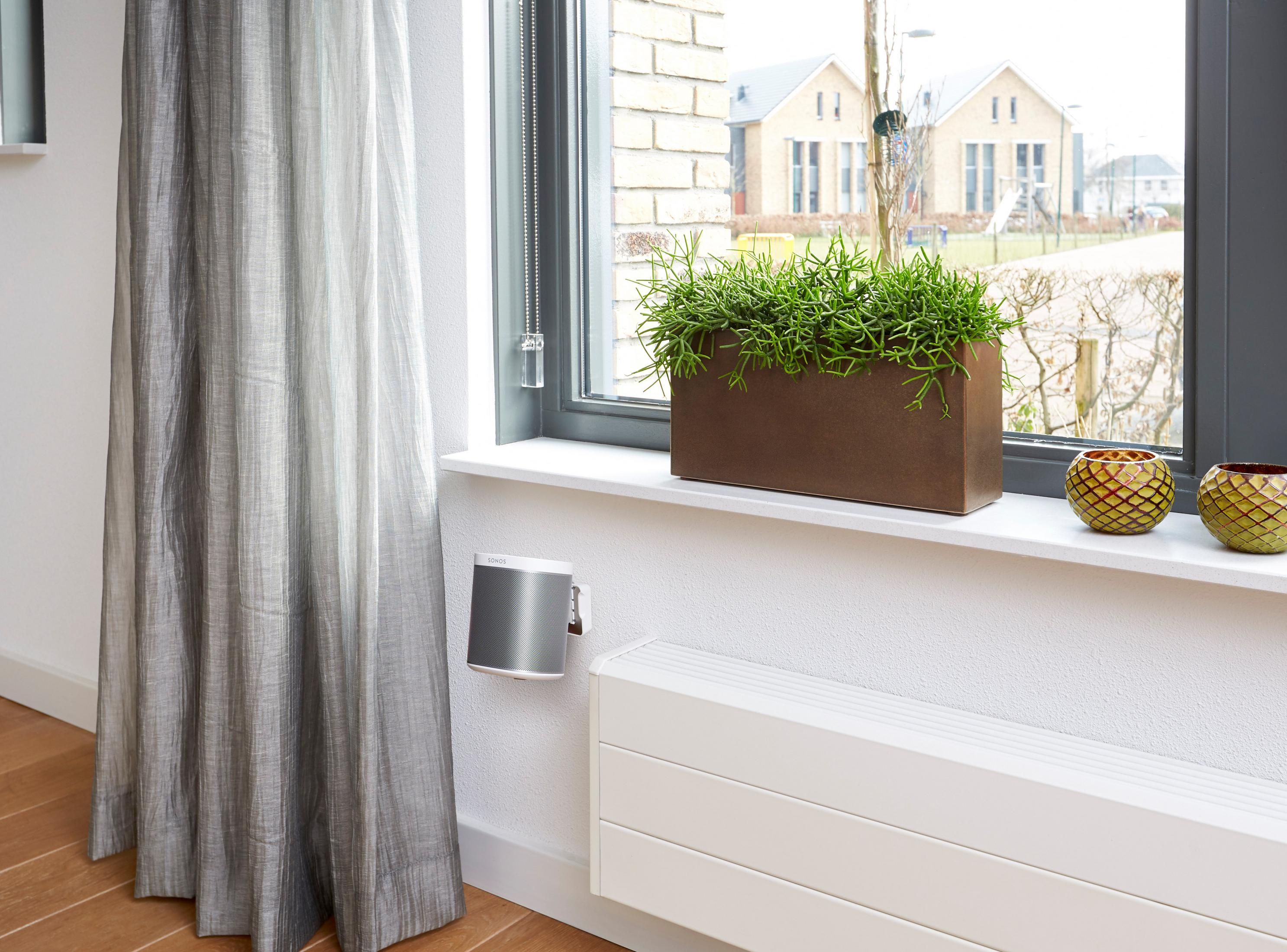 Interior de la habitación con ventana, cortina, planta y altavoz en la pared.