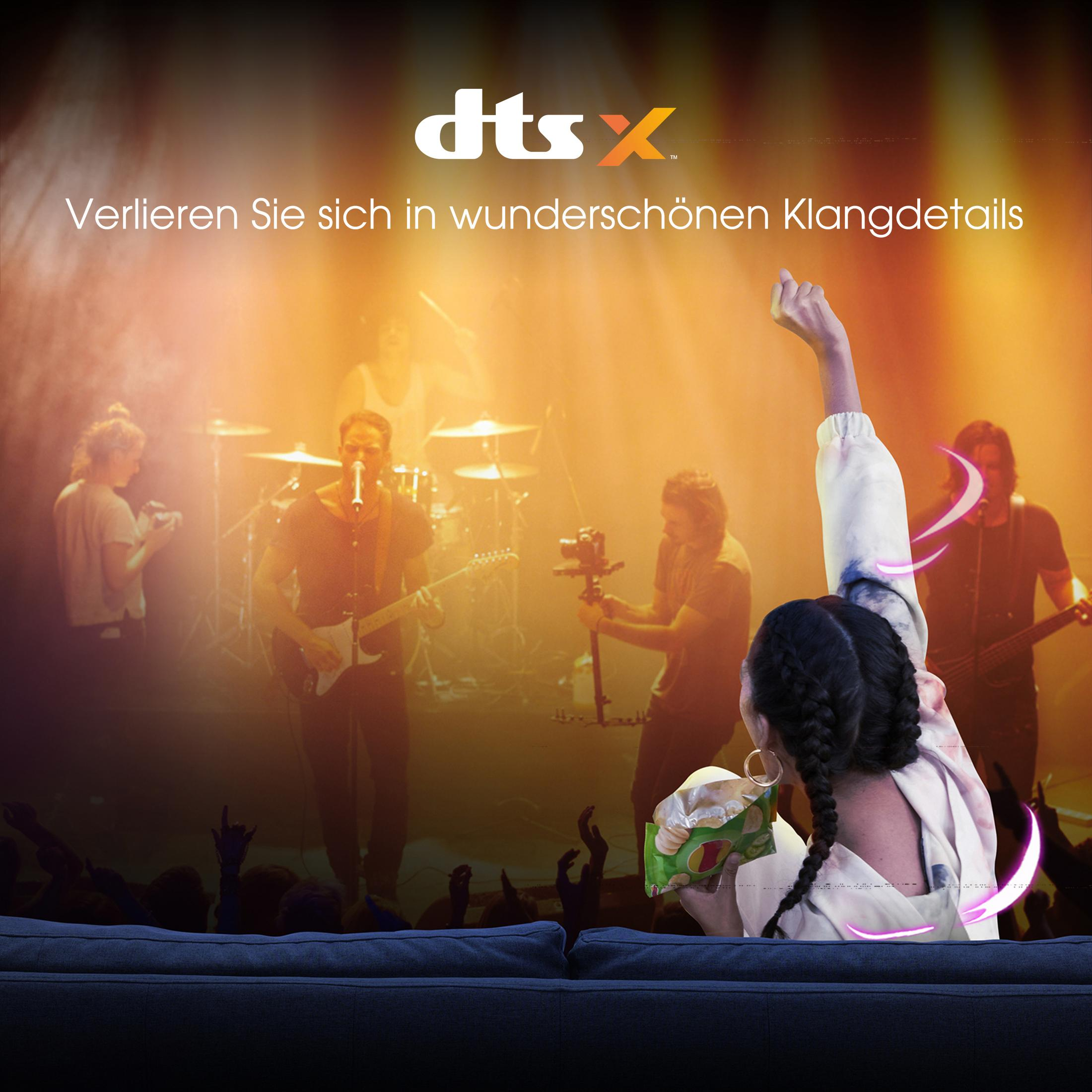 Publiczność na koncercie, kobieta trzyma przekąskę. Logo DTS:X w lewym górnym rogu.