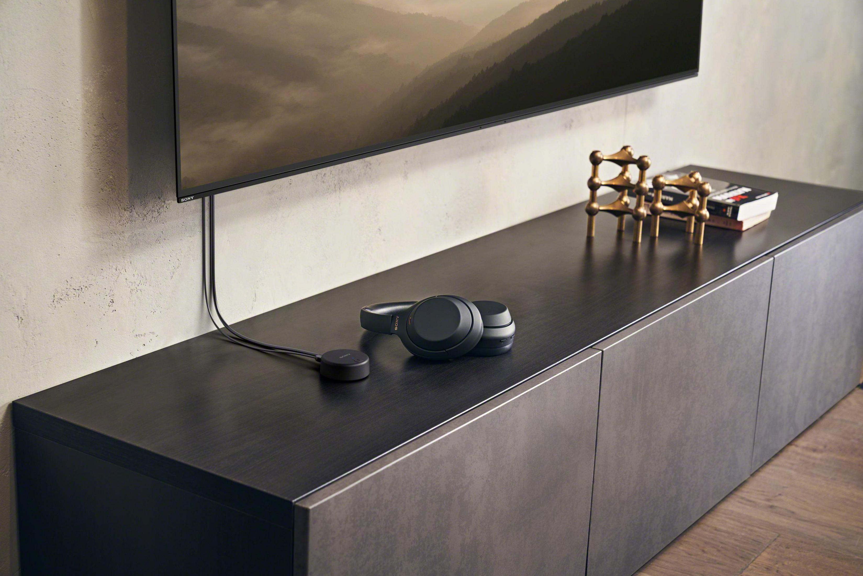 Auriculares y un mando a distancia sobre un mueble de madera oscura. Un televisor en el fondo.