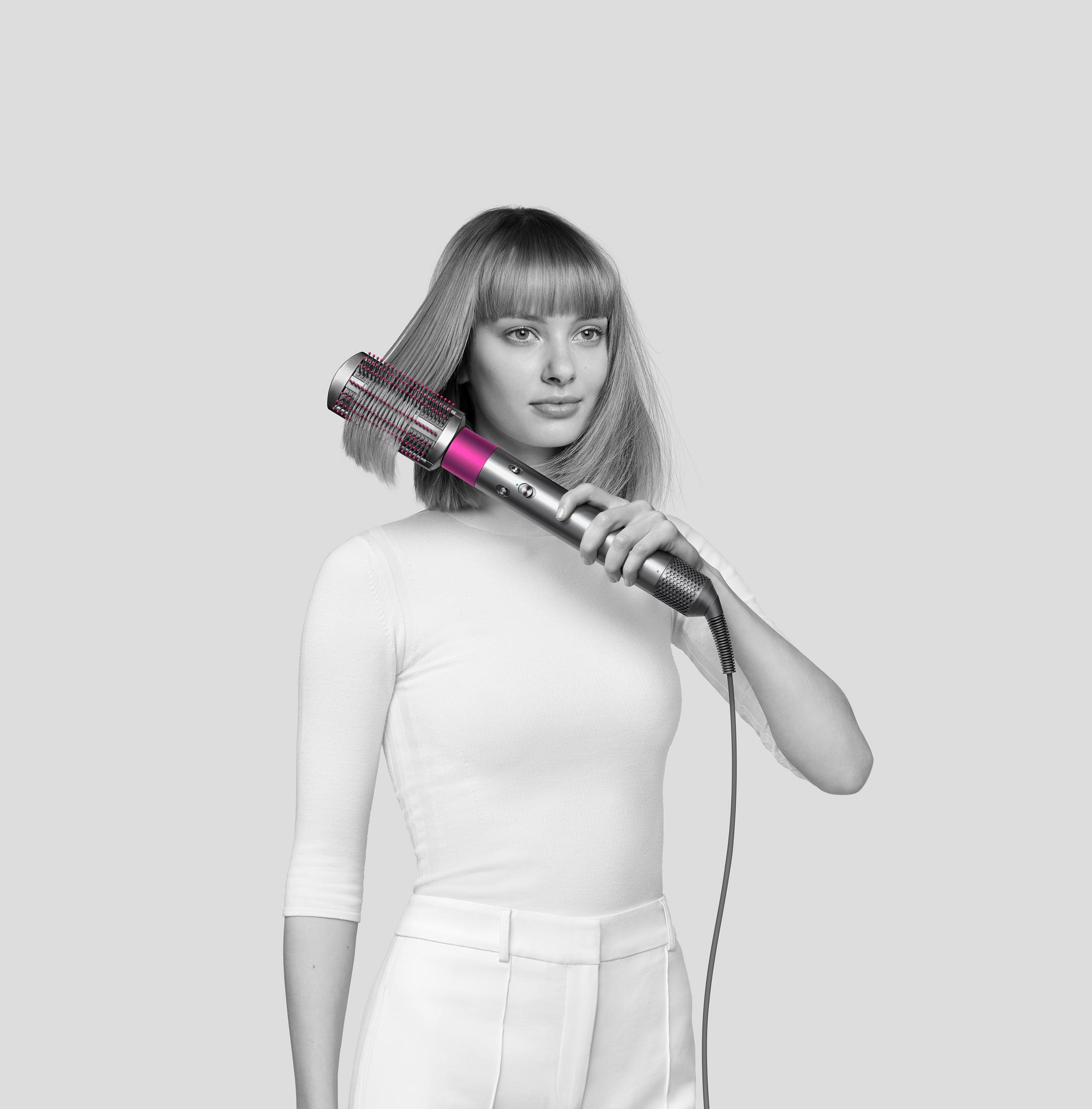 Frau benutzt ein rosa-silbernes Haarstyling-Gerät. Sie trägt weiße Kleidung, kurzes blondes Haar.