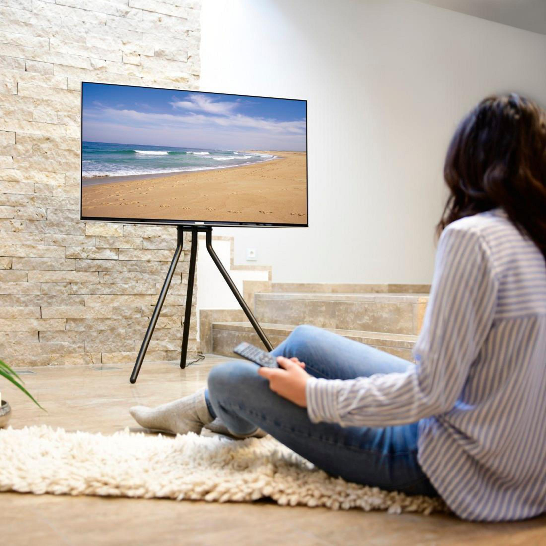 Kobieta siedzi na dywanie, oglądając telewizję na stojaku. Scena plażowa na ekranie.