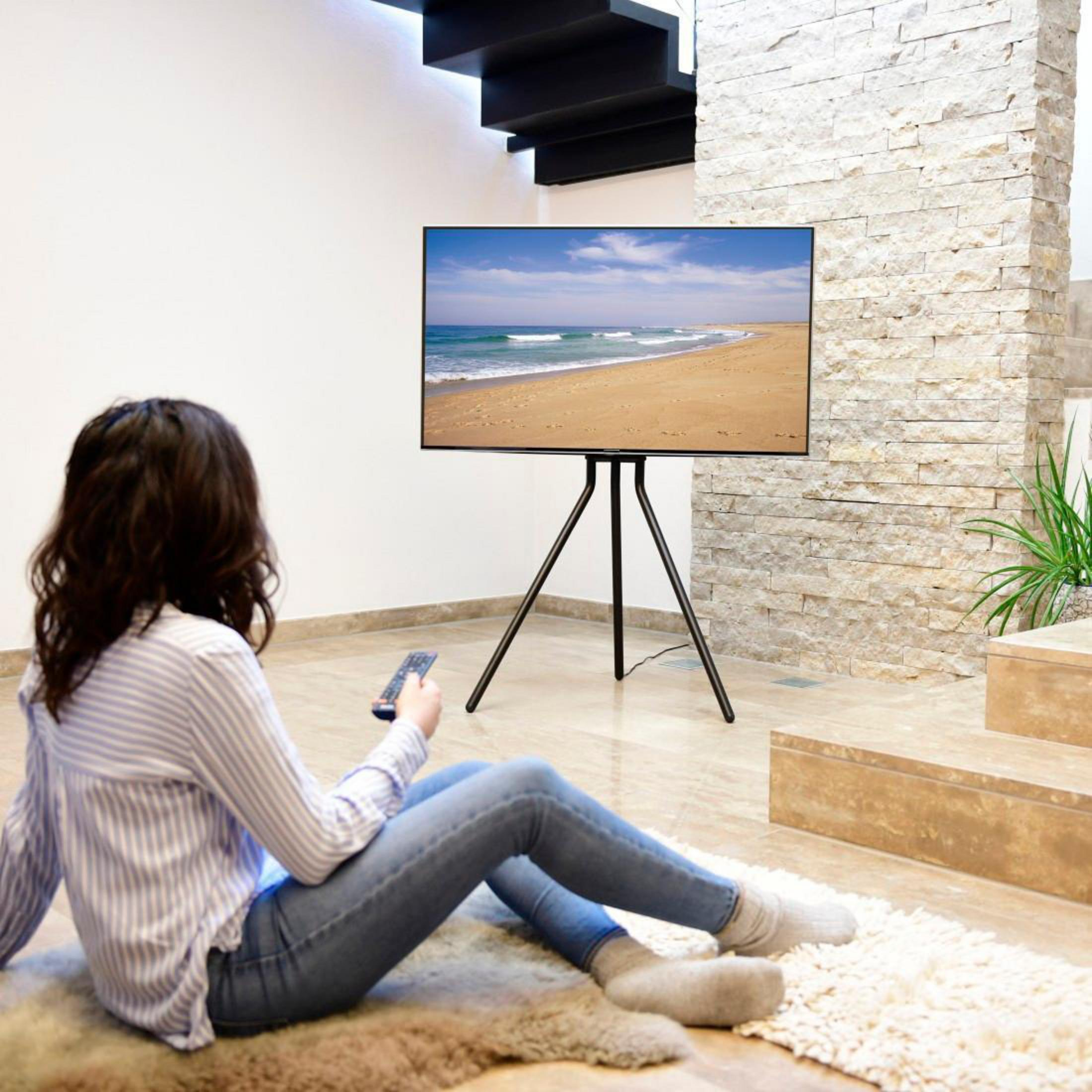 Kobieta siedzi na podłodze, oglądając telewizję na stojaku. Telewizor pokazuje scenę z plaży.