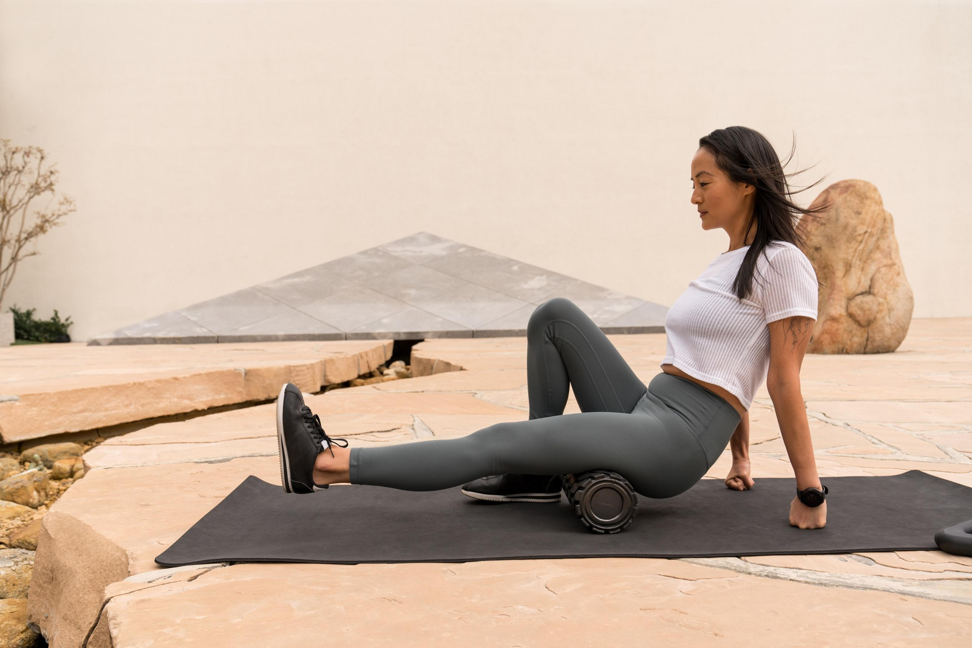 Mujer en una esterilla haciendo ejercicio de piernas con un rodillo de espuma. Ropa deportiva, al aire libre.