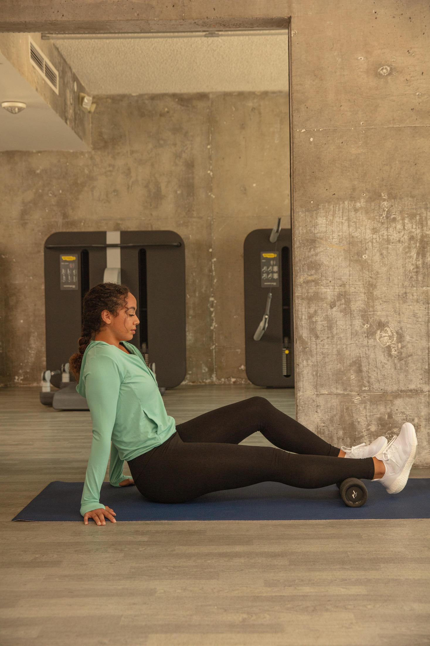Eine Frau dehnt ihr Bein auf einer blauen Matte mit einer Faszienrolle im Fitnessstudio.