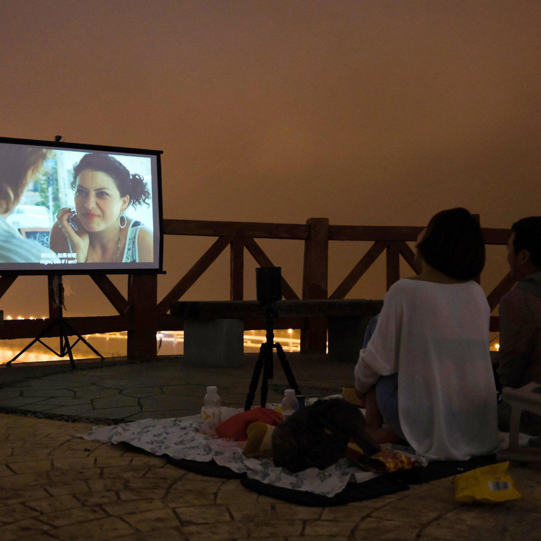 Leute schauen sich abends im Freien einen Film an, der auf eine Leinwand projiziert wird. Es gibt ein Picknick.