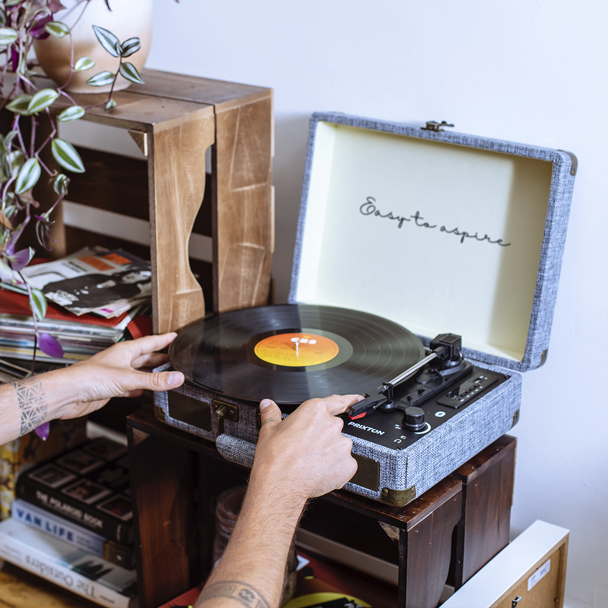 Ręce umieszczają płytę winylową na gramofonie. Drewniane skrzynie i rośliny w tle.
