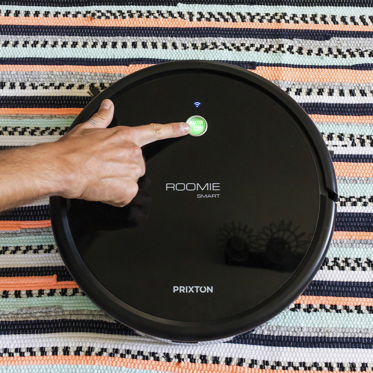 Czarny robot odkurzający z ręką naciskającą przycisk 'auto' na kolorowym dywanie.