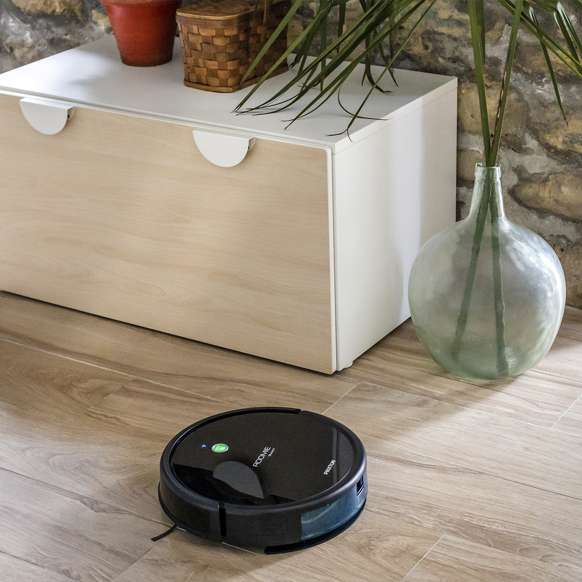 Czarny robot odkurzający na drewnianej podłodze. W tle szafka i wazon.