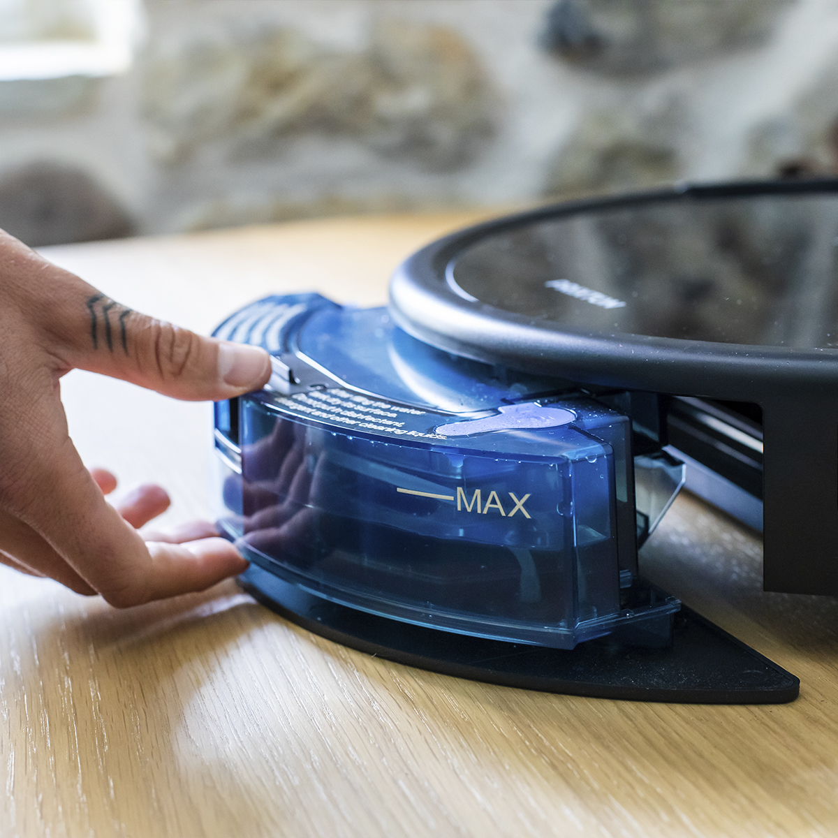 Ręka osoby obsługuje niebieski zbiornik na wodę czarnego robota odkurzacza. Poziom max jest oznaczony.